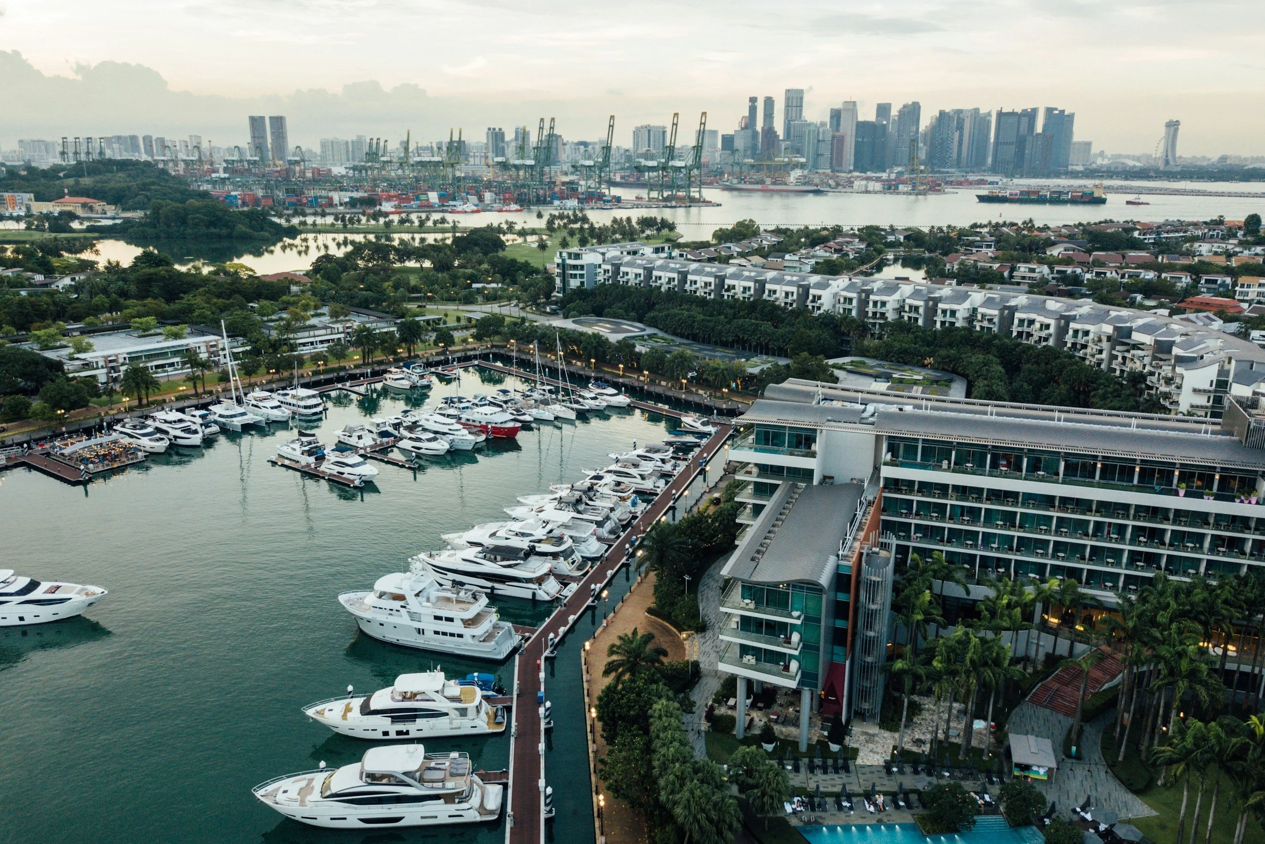 An aerial view of a marina filled with yachts and boats, surrounded by modern buildings, with a city skyline and harbor in the background.