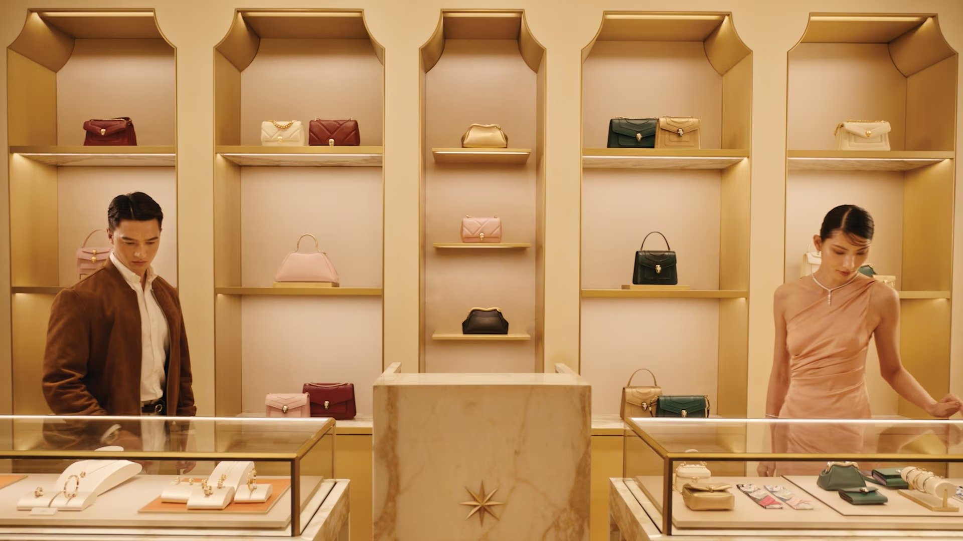 Inside a luxury boutique jewelry store with gold accents, featuring display cases with jewelry and handbags, and two models shopping.