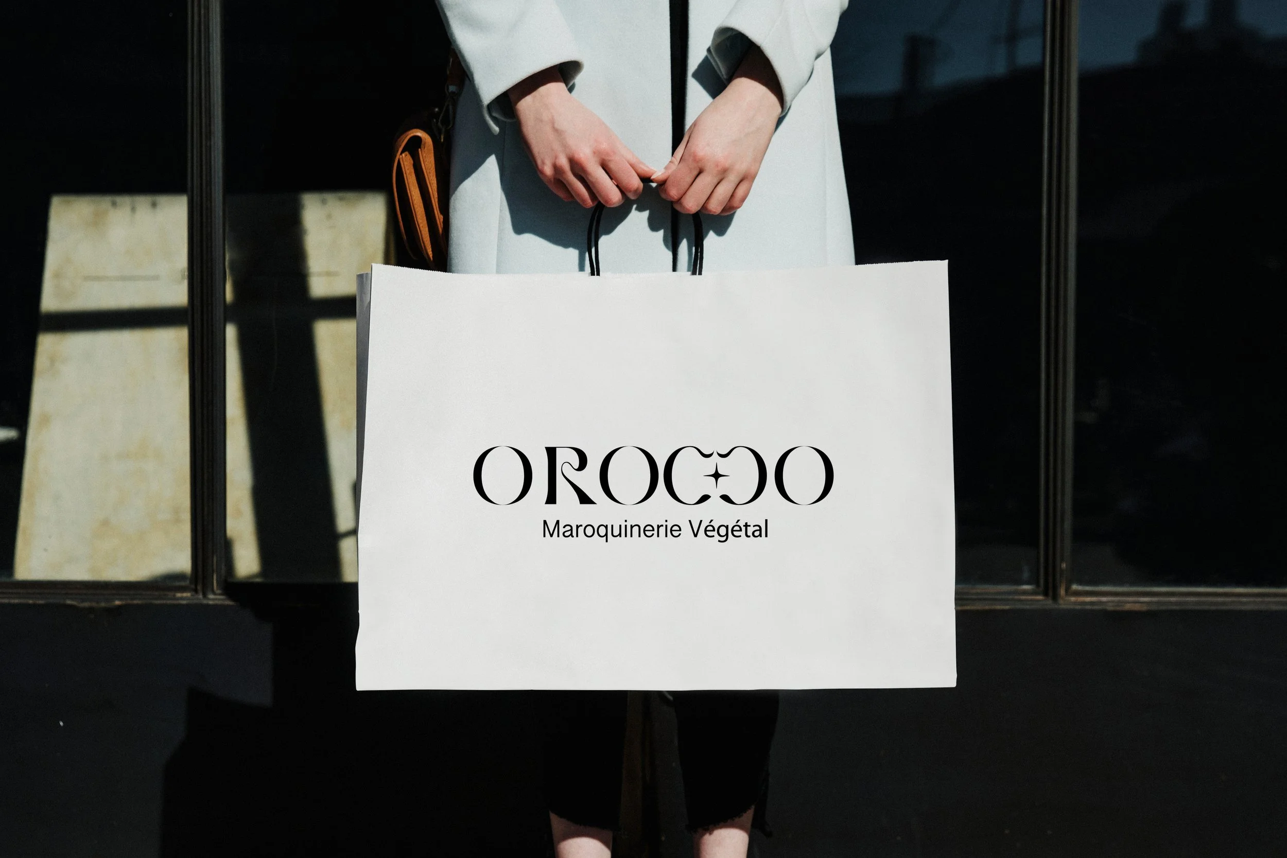 Une personne tenant un grand sac blanc avec le logo 'OROCO' et le texte 'Maroquinerie Végétal', devant une façade en verre.