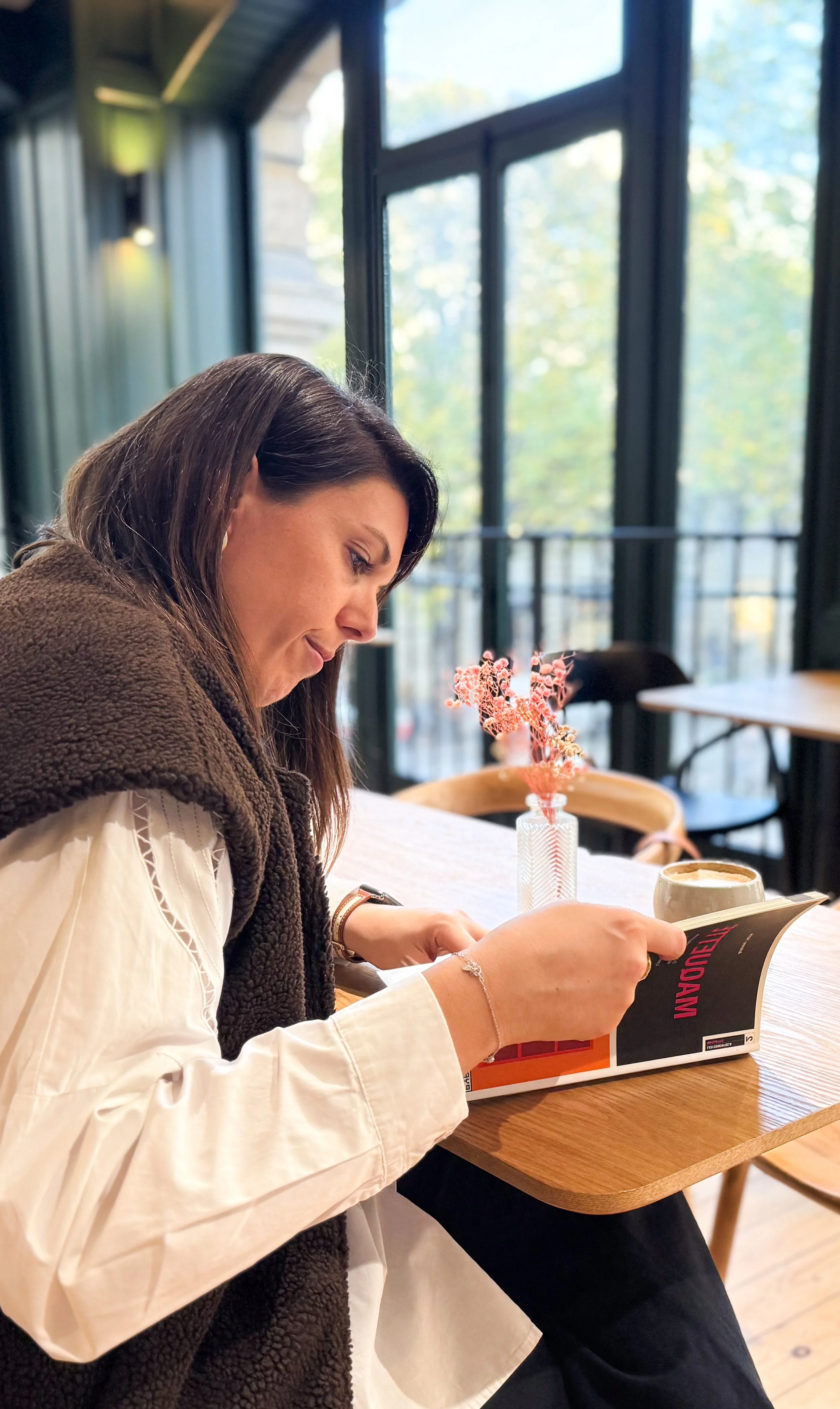 Femme lisant un magazine dans un café avec une tasse de café et une petite plante en vase sur la table.