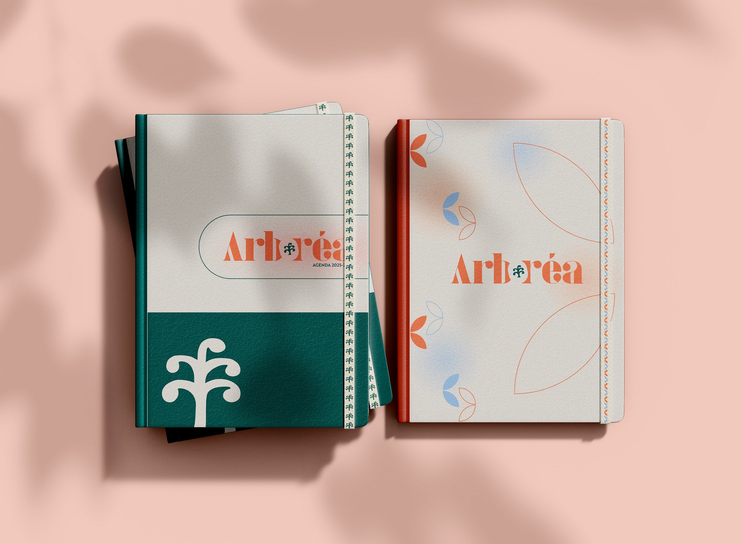 Deux agendas pour 2023 avec le mot 'Arbrea' en orange, un à gauche avec une couverture verte et blanche, et un à droite avec une couverture blanche et des motifs de feuilles en rouge et bleu, posés sur une surface rose clair.