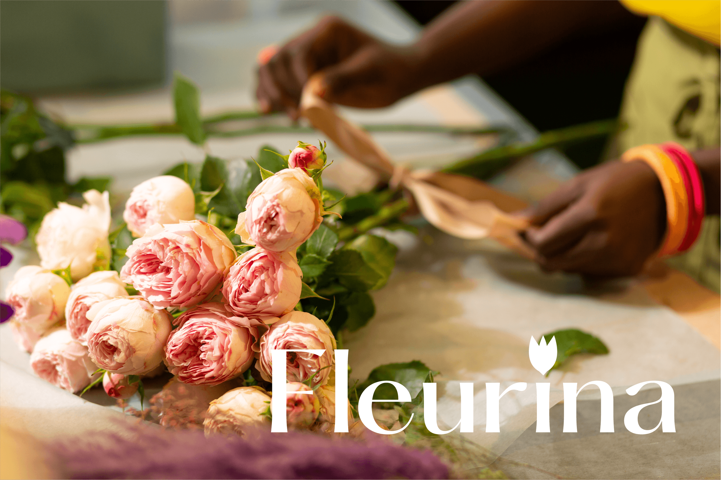 Un bouquet de roses roses avec une personne en arrière-plan en train de faire une composition florale. Le mot "Fleurna" apparaît en bas de l'image.