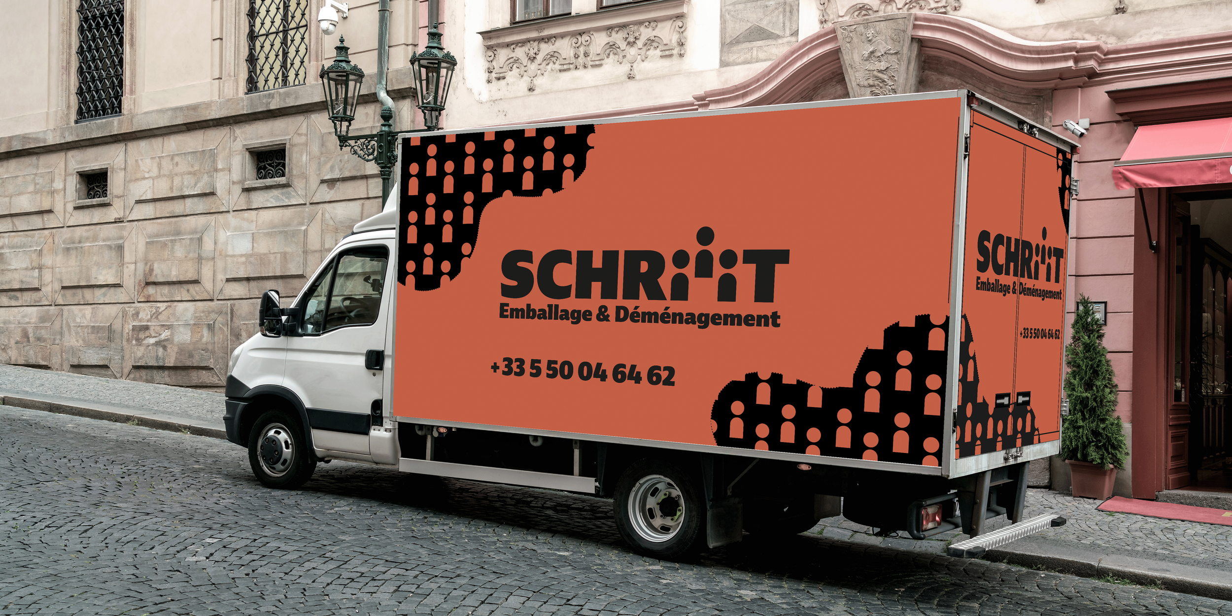 Camion blanc avec une publicité orange pour une entreprise d'emballage et déménagement, affichant le nom SCHRiTT et un numéro de téléphone, stationné sur une rue pavée, avec un bâtiment en pierre en arrière-plan.