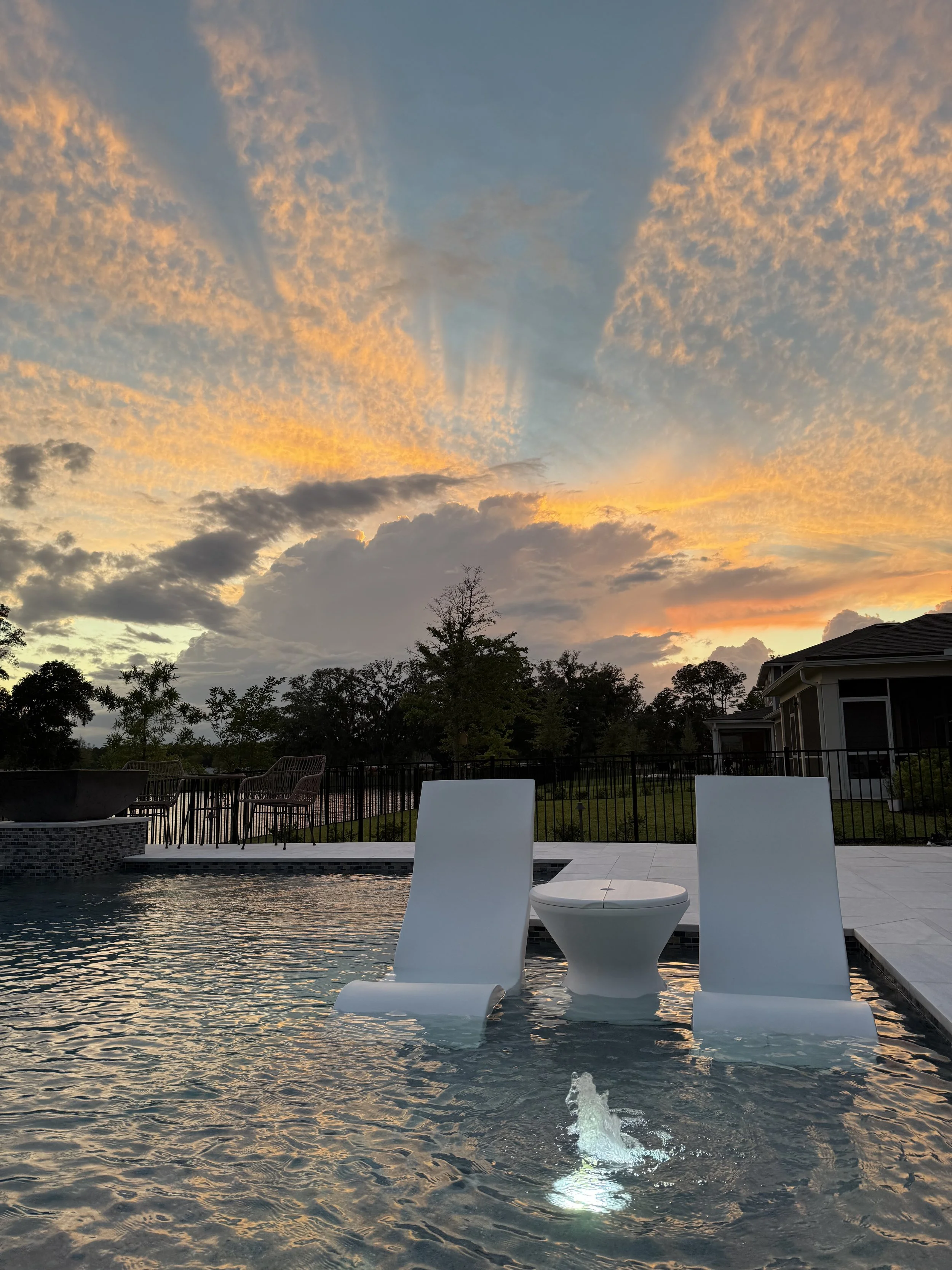 Poolside scene with two white lounge chairs partially submerged in water and a small round table between them, sunset sky with colorful clouds and trees in the background.