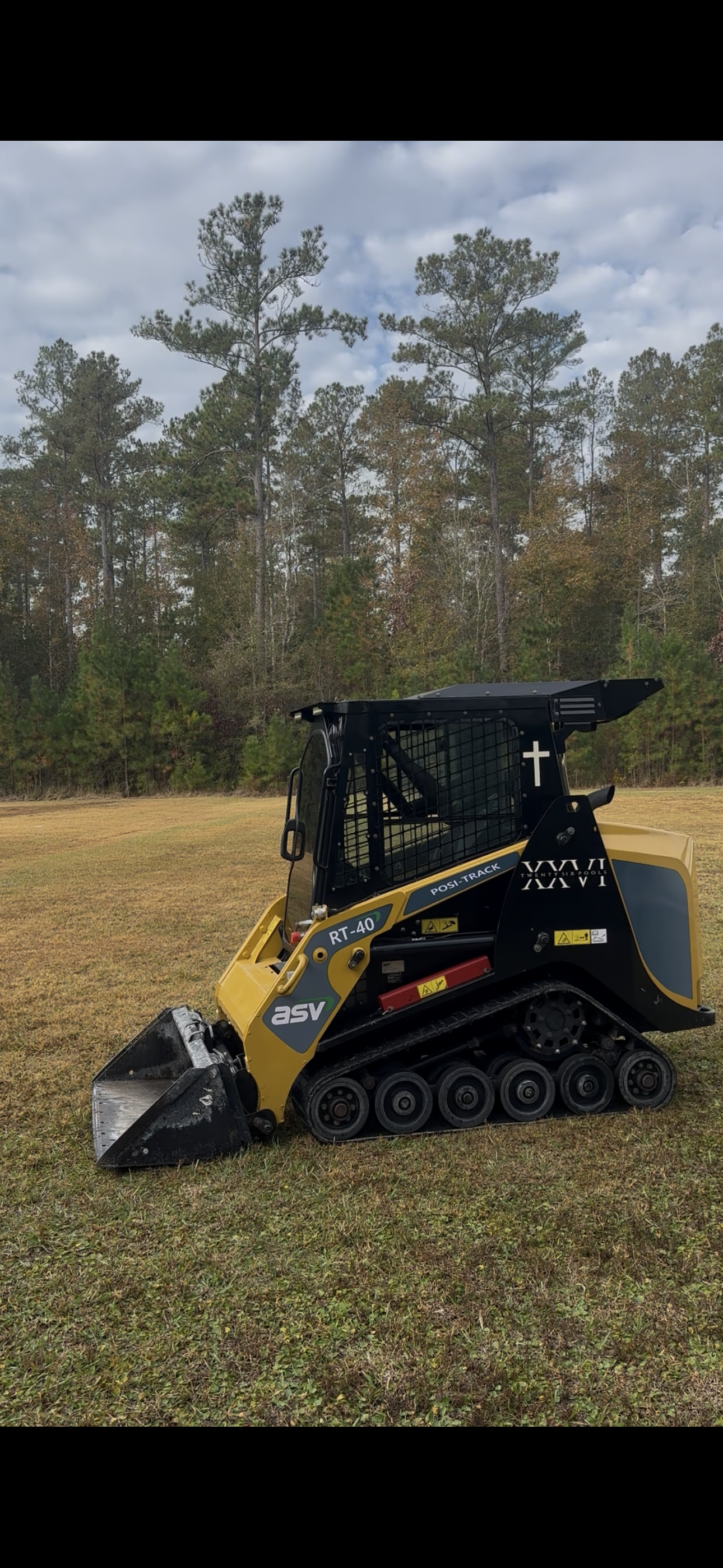 A yellow and black tracked skid steer loader on grass, with trees and a cloudy sky in the background. Kingsland GA.