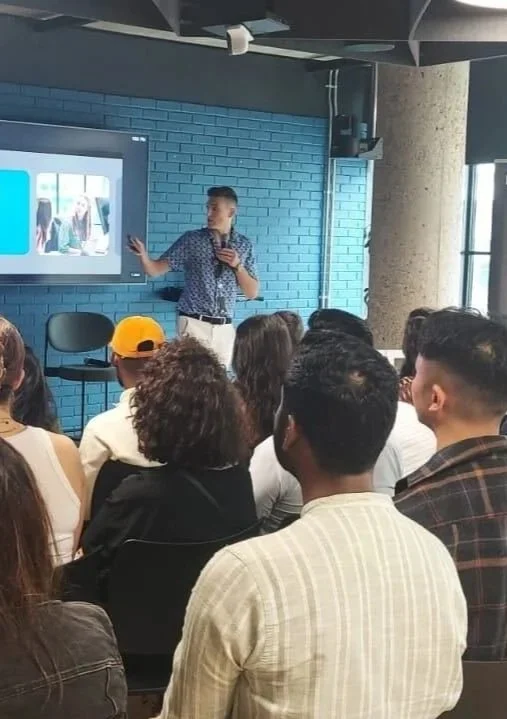 A young man giving a presentation to a group of diverse people in a modern room with a blue brick wall and a large screen.
