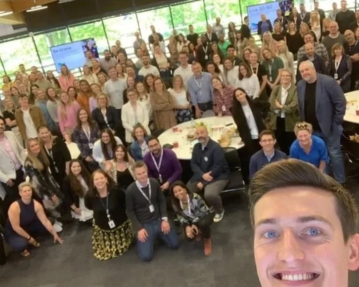 Group of diverse professionals at a conference or workplace event, smiling and posing for a selfie in a modern office setting with large windows and digital screens in the background. The reference man is taking the picture.