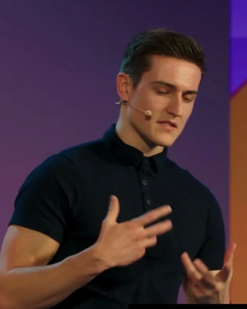 The reference man. A young man with dark hair and light skin is wearing a black polo shirt and using a headset microphone. He appears to be mid-speech or presentation on a stage with purple and orange background lighting.