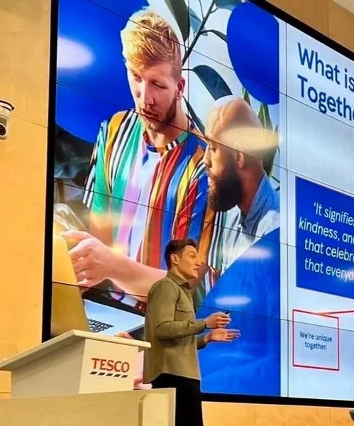 A man is giving a presentation in front of a large digital screen displaying an image of two young men looking at a smartphone. The screen shows text about 'we're unique together' and includes a blue and white background in a conference room.