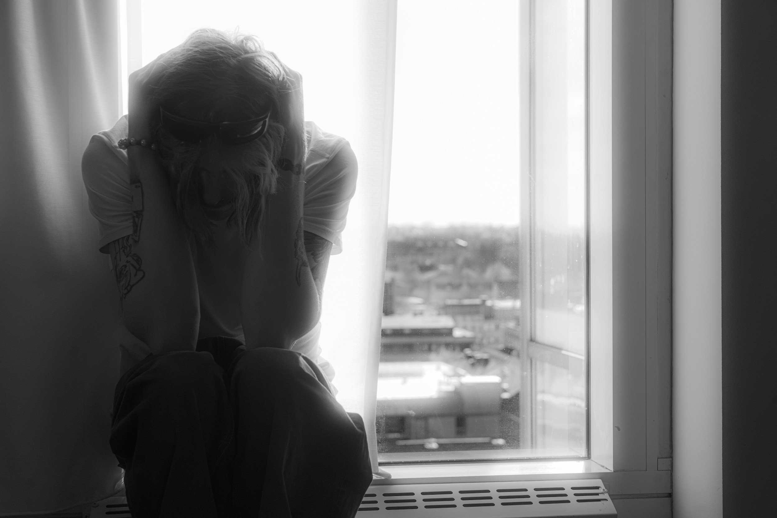 A person with long hair sitting by a window with blinds, looking down with their head resting on their hand. The scene is black and white.