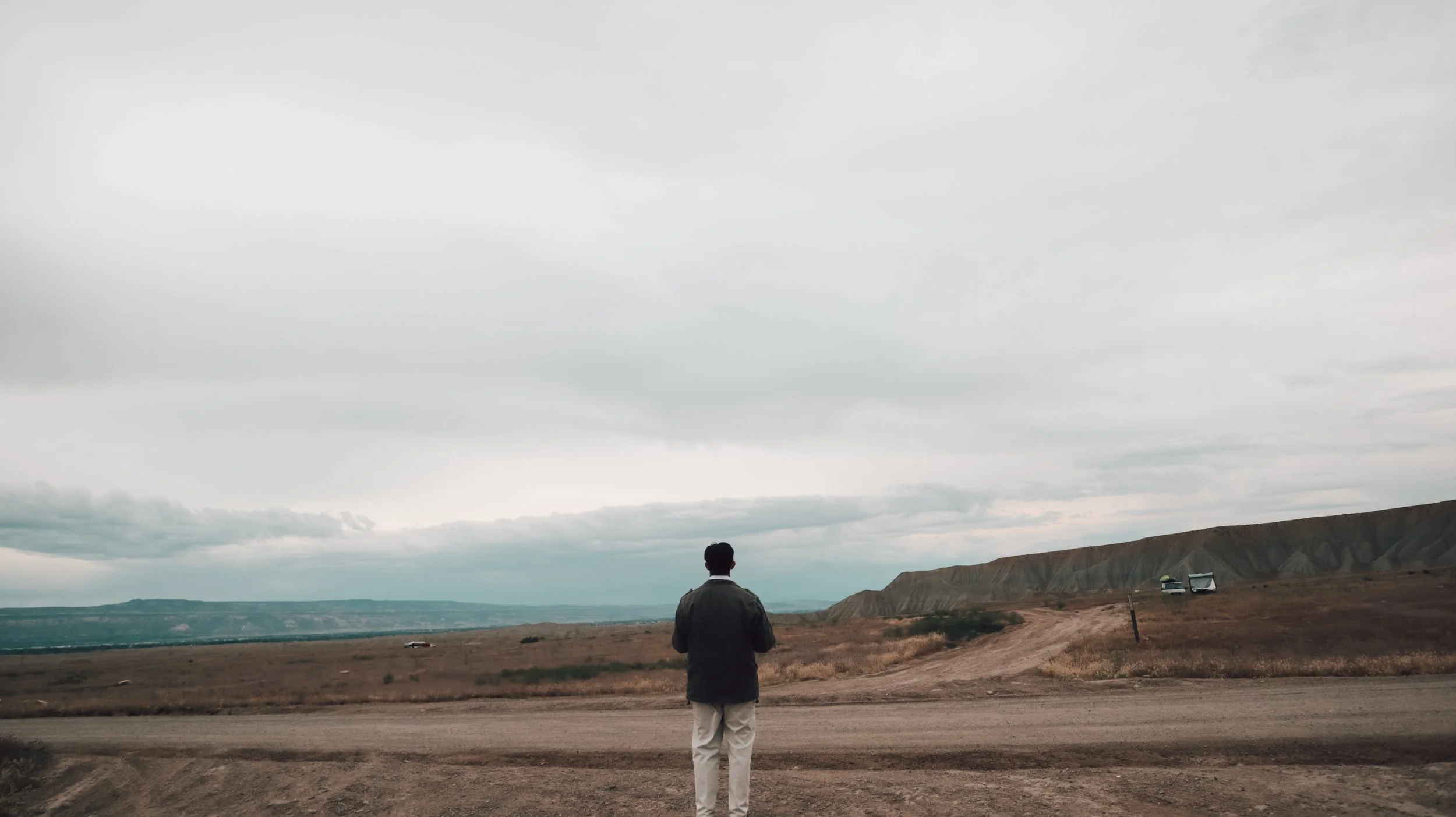 A person stands in an open landscape with a mountain range in the distance, overcast sky, and a few vehicles nearby.