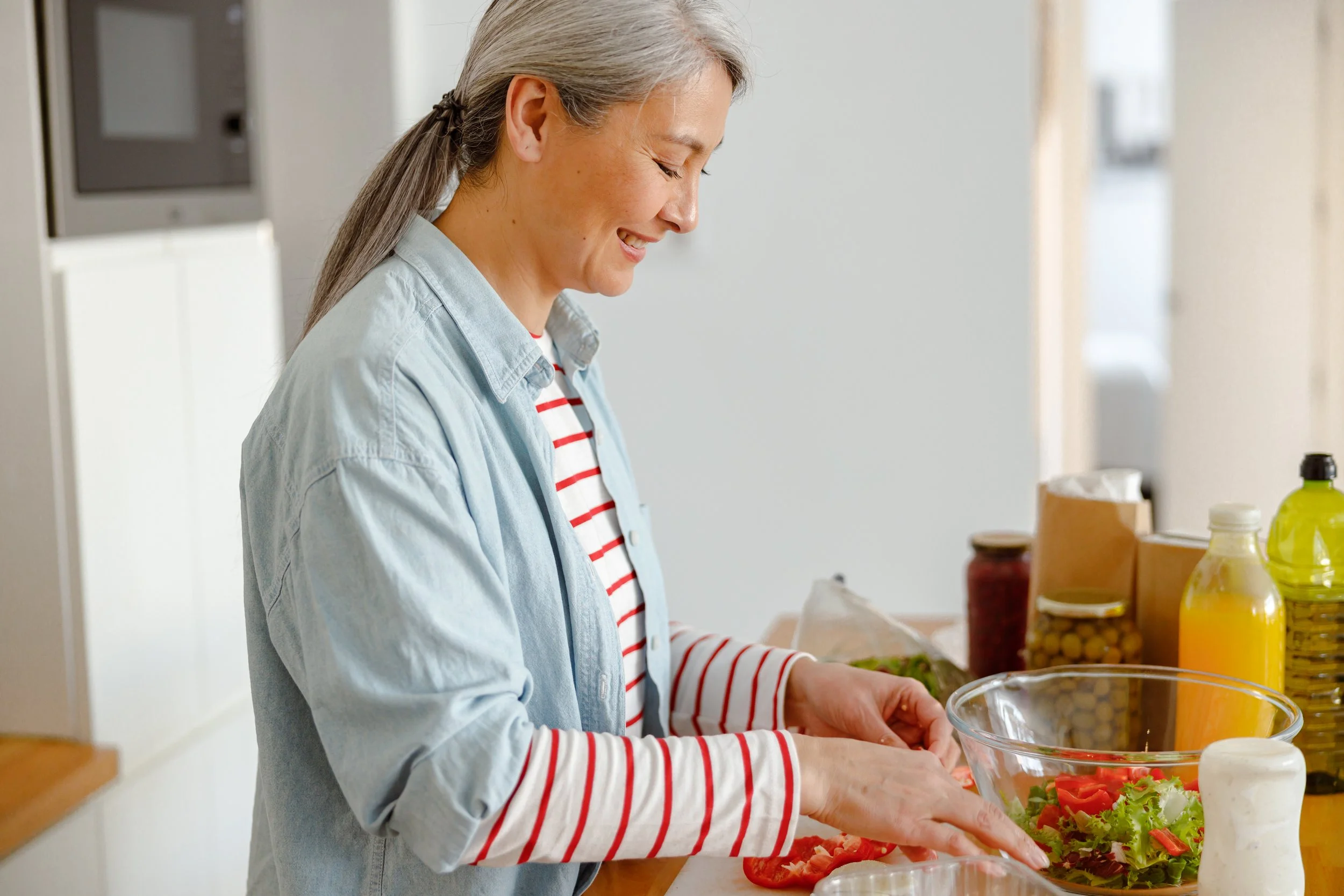 A woman with gray hair in a ponytail, wearing a light denim shirt over a red and white striped long sleeve shirt, preparing a salad in a bright kitchen.