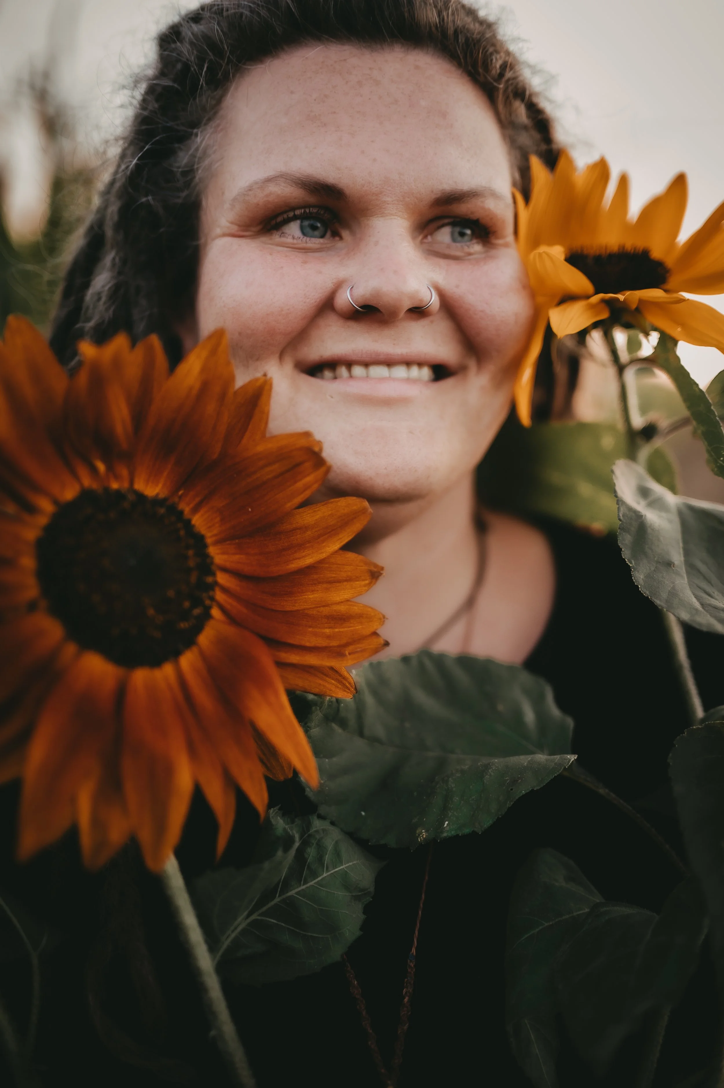 Ein lachendes Mädchen mit blauen Augen und Nasenring umgeben von Sonnenblumen im Freien.