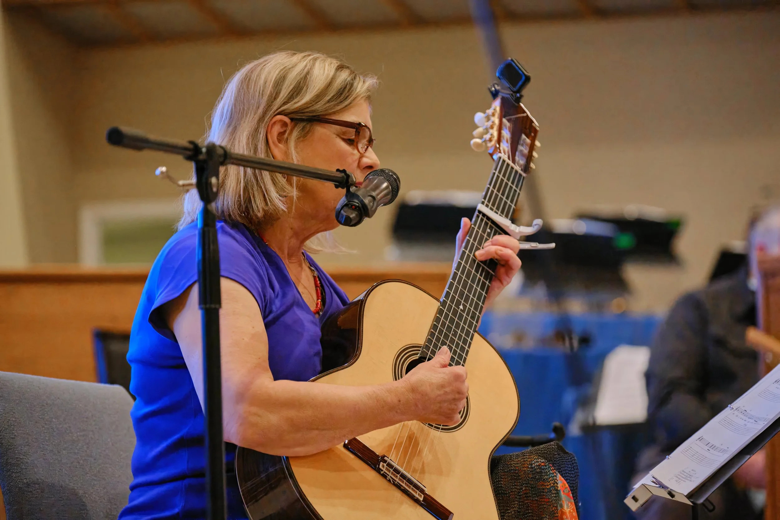 A woman with glasses and gray hair plays an acoustic guitar in a music rehearsal space, singing into a microphone.