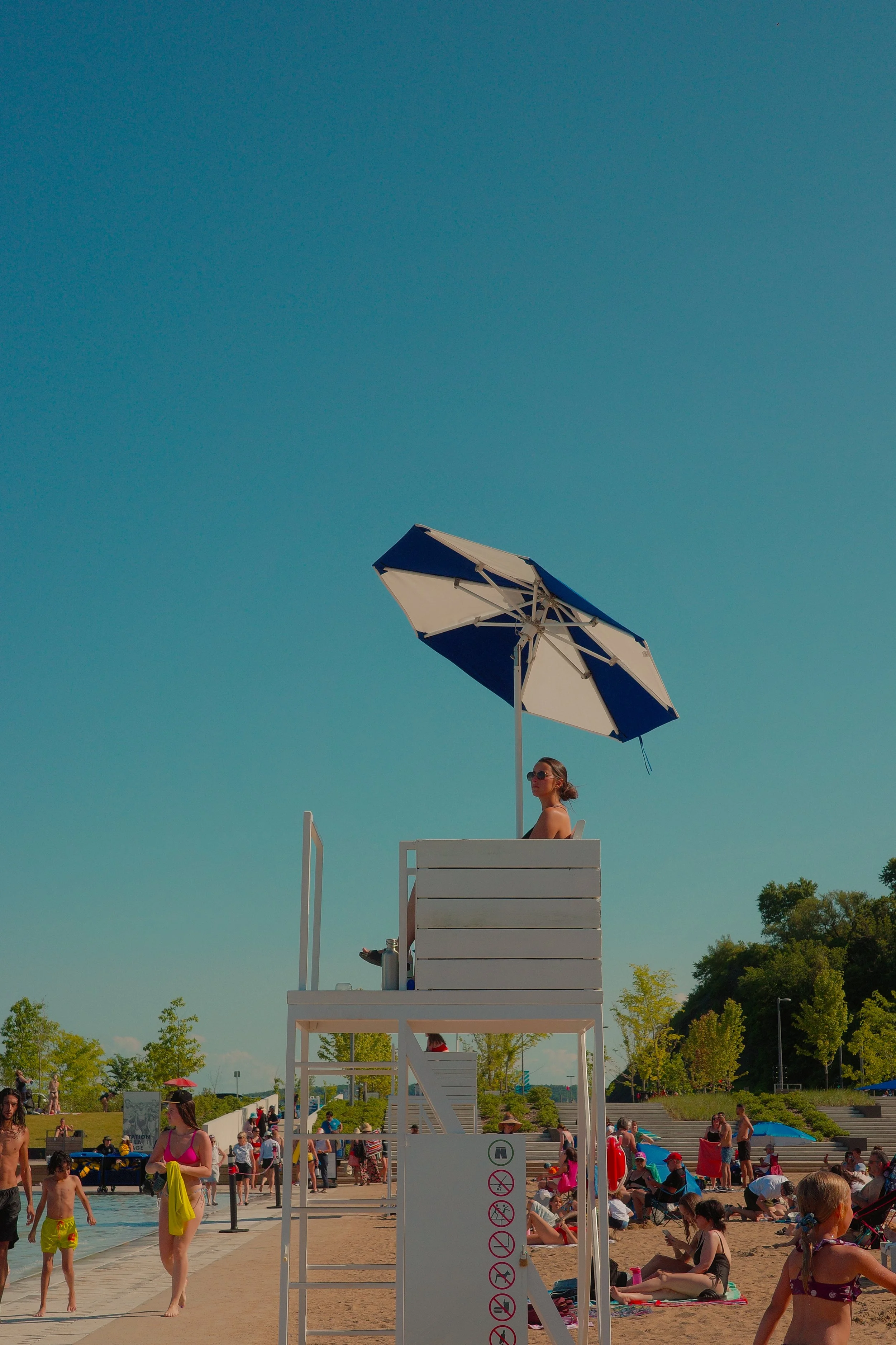Une surveillante de baignade en position de garde sur une tour en haut d'une plage bondée, sous un parasol. Des gens se baignent et se détendent sur la plage par une journée ensoleillée.