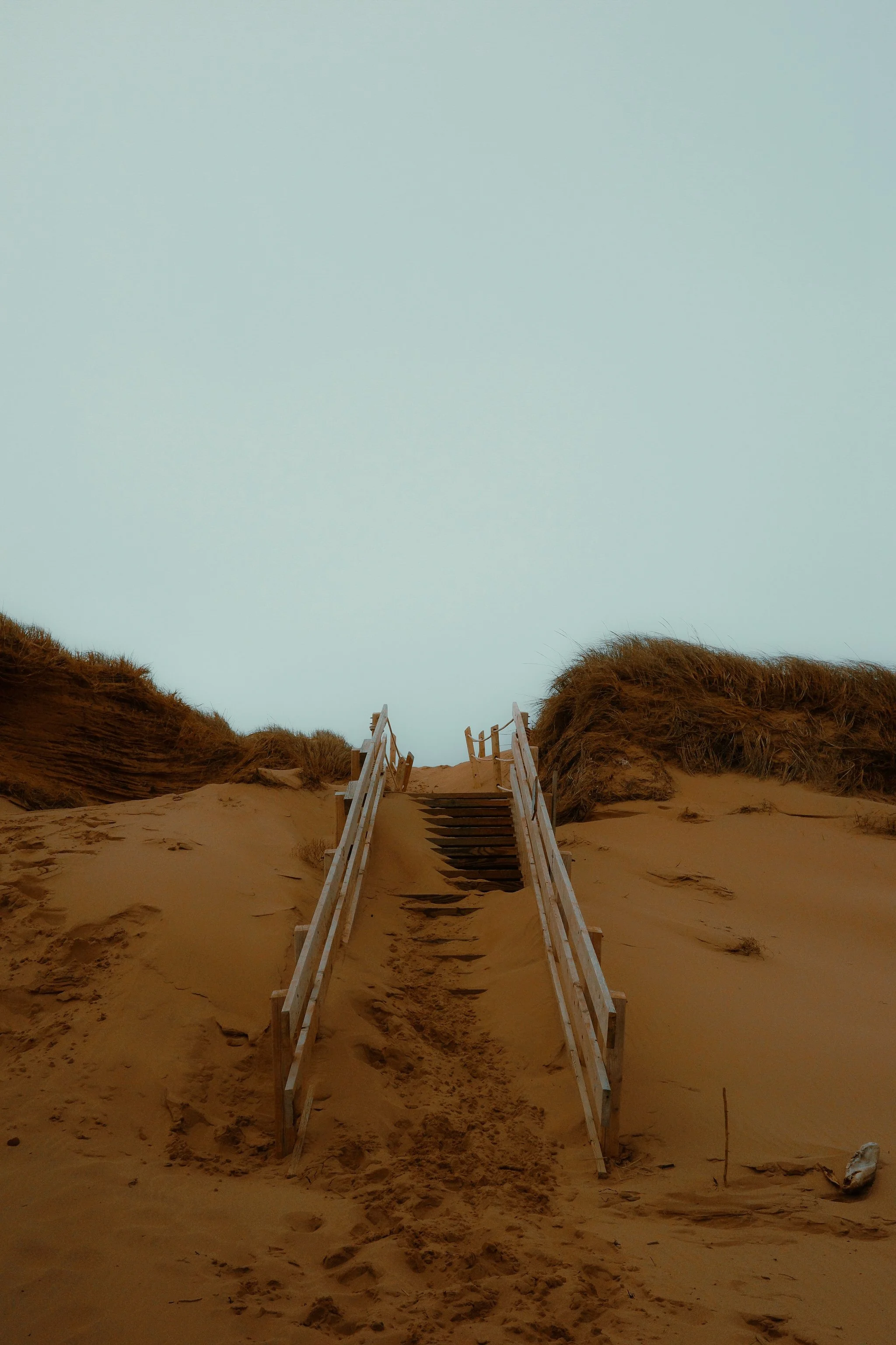 Une passerelle en bois reliant une dune de sable à une zone plus élevée, avec un ciel clair en arrière-plan.