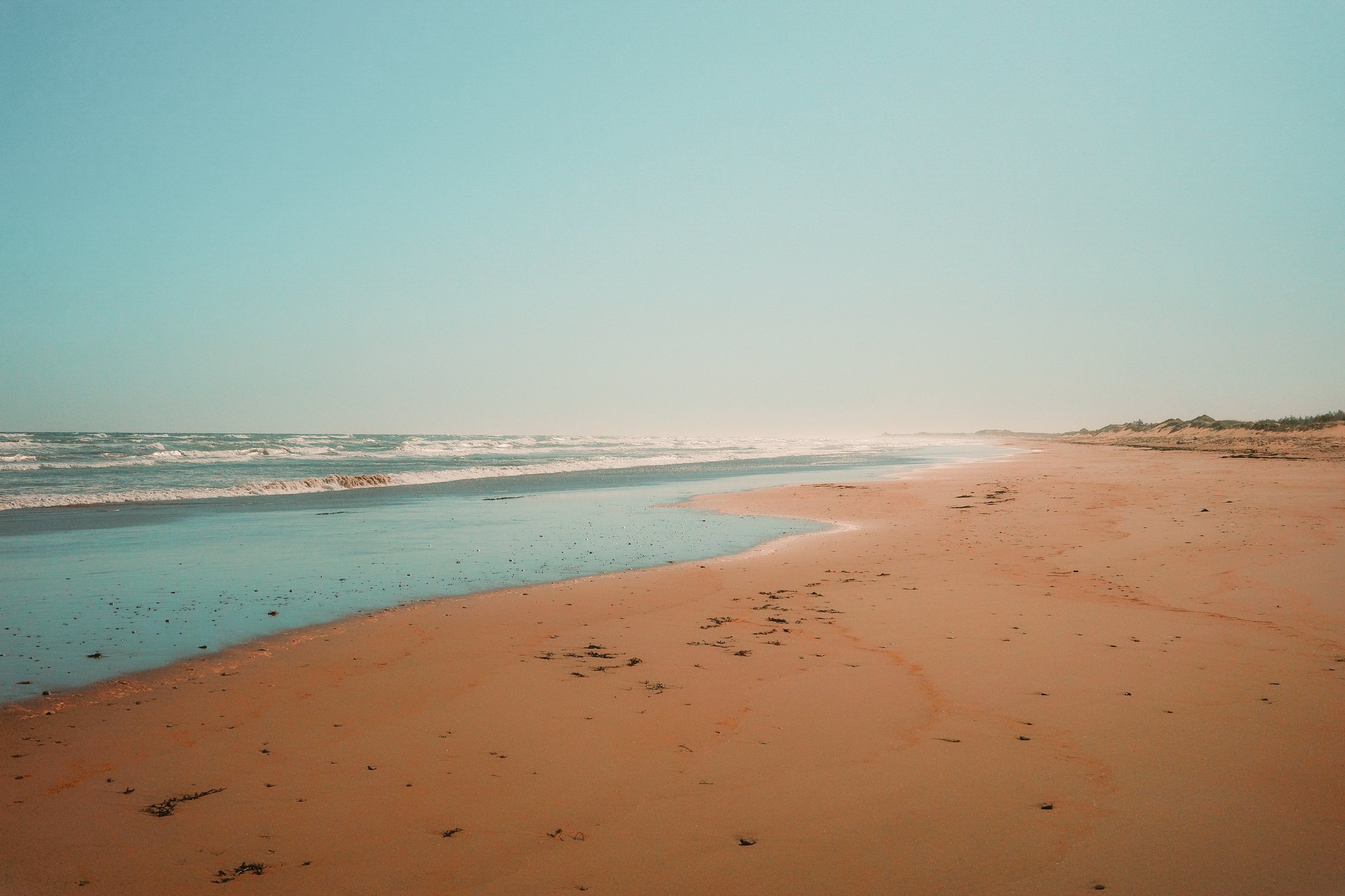 Plage de sable avec eaux calmes et vagues, ciel clair et horizon lointain.