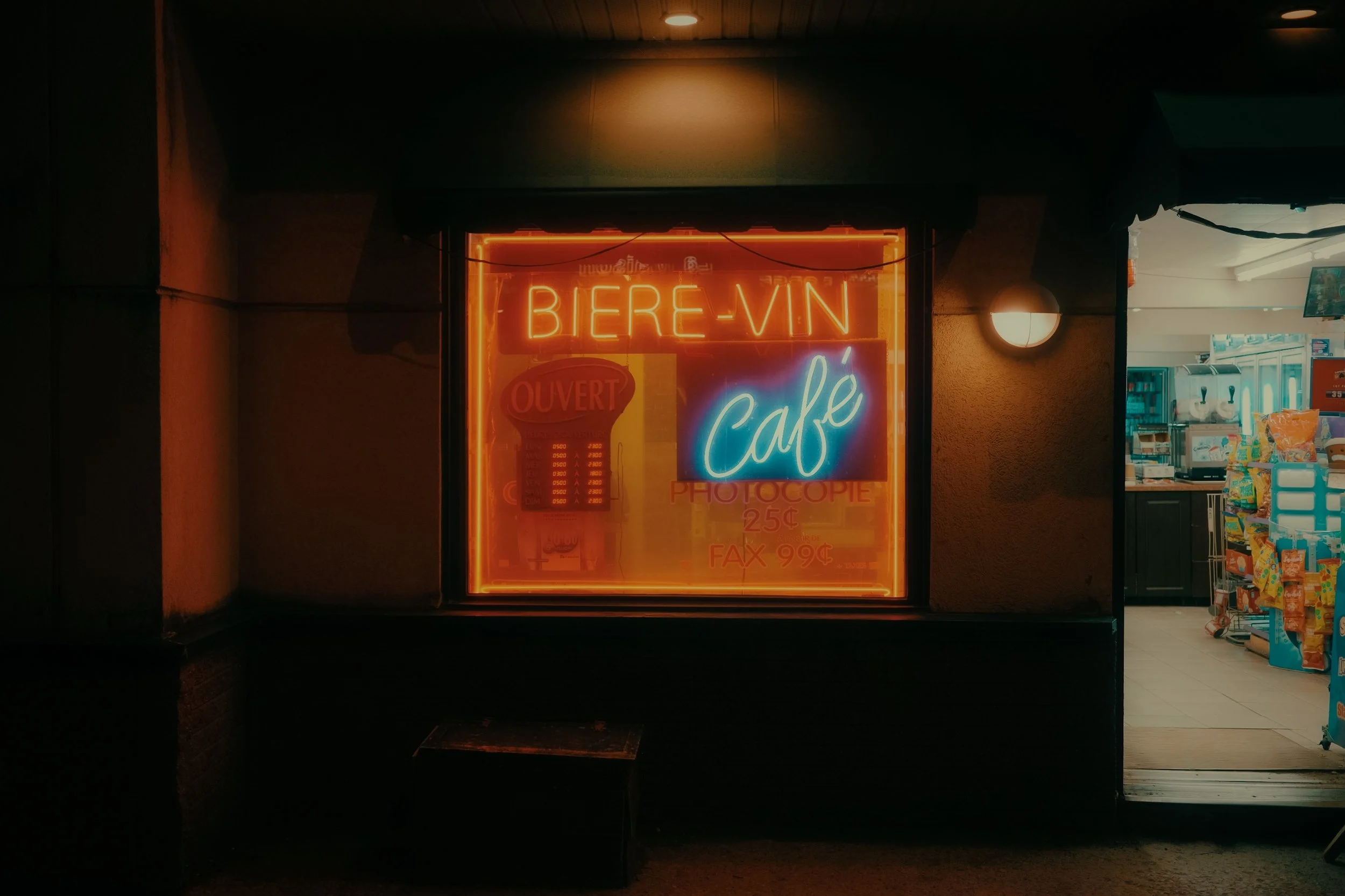 Une vitrine d'un café avec des néons orange et bleus affichant 'BIERE-VIN' et 'Café', à l'intérieur on voit des produits de konbini.