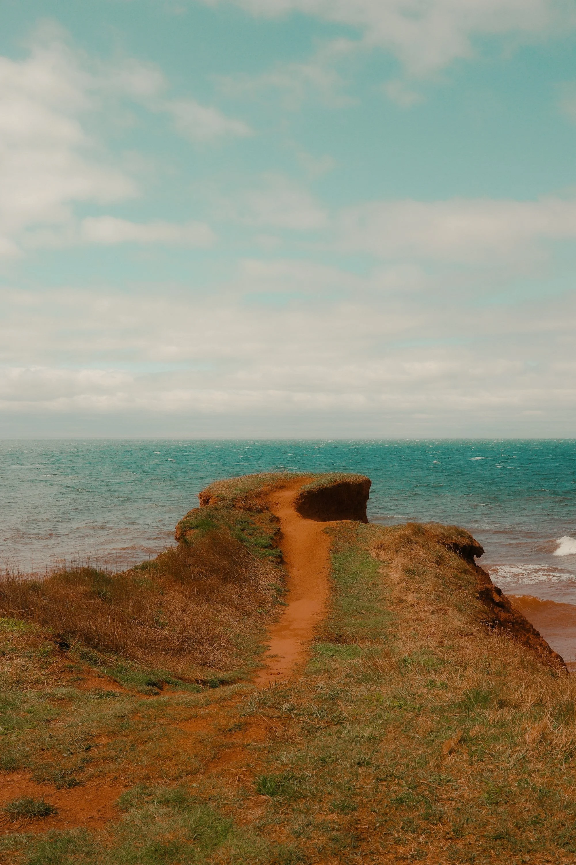 Sentier de terre rouge conduisant à une falaise au bord de la mer, avec un ciel nuageux au-dessus.