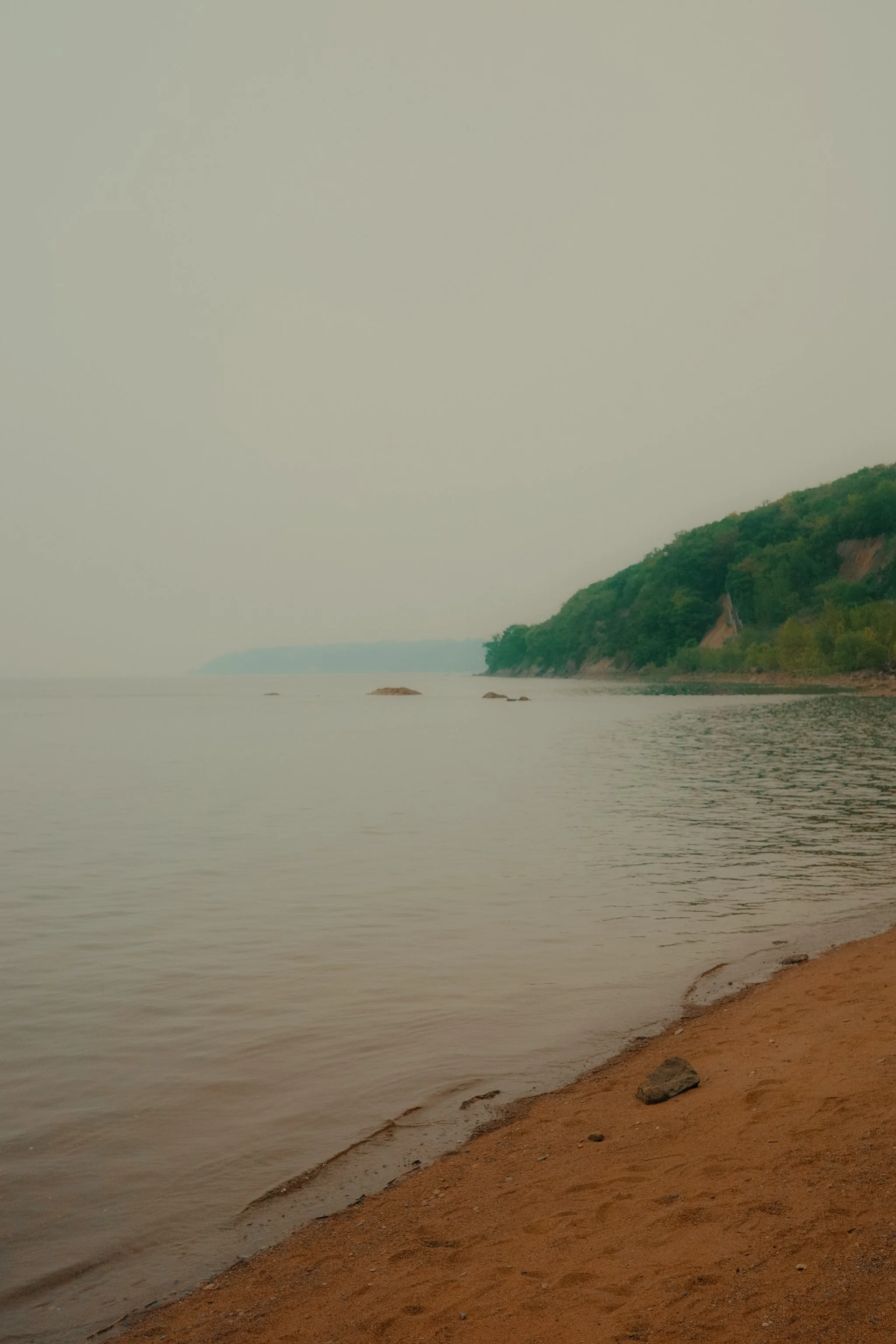Plage de sable avec des rochers, eaux calmes et une côte boisée sous un ciel nuageux.