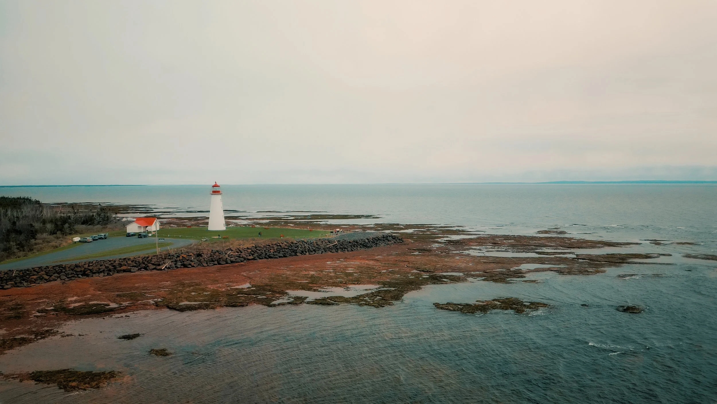 Phare blanc avec une toiture rouge, situé sur une côte rocheuse, face à la mer, avec un petit bâtiment et des voitures dans une zone herbeuse adjacente.