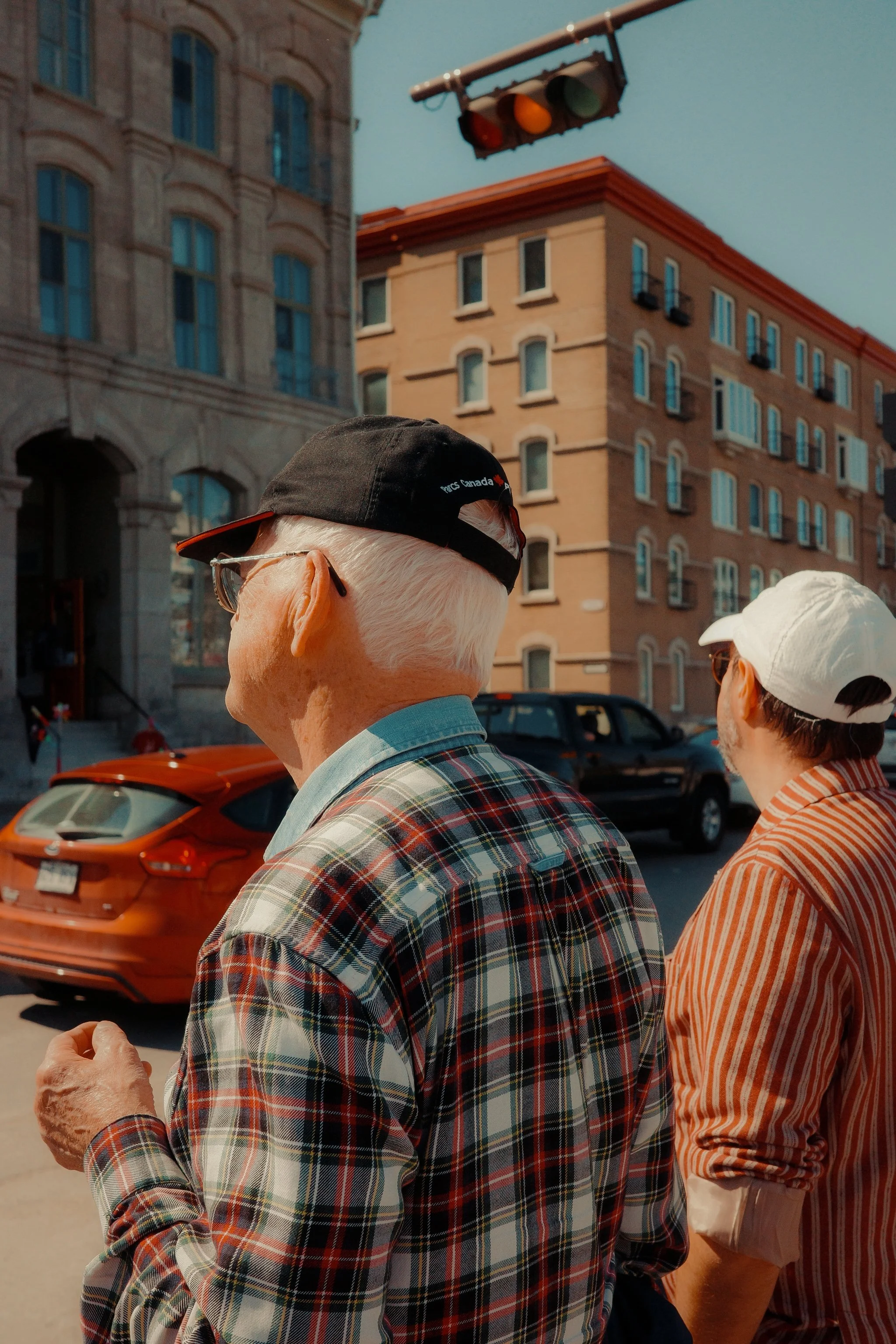 Deux hommes âgés marchant dans une rue urbaine, portant des casquettes, avec des bâtiments en arrière-plan et une lumière de feu de circulation.