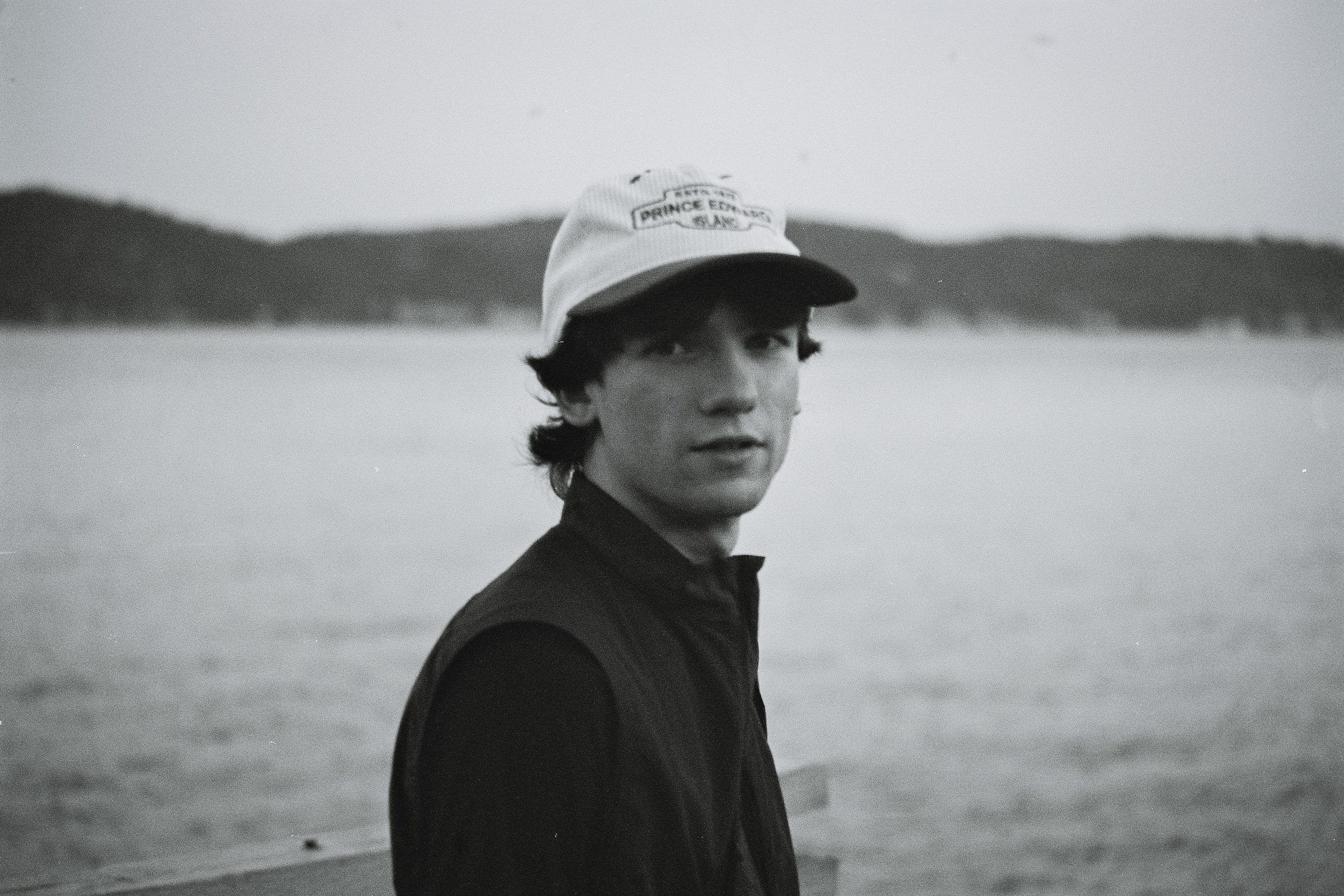 Un jeune homme avec des cheveux ondulés portant une casquette blanche et un vêtement sombre, se tenant près d'un plan d'eau avec des montagnes en arrière-plan, en noir et blanc.