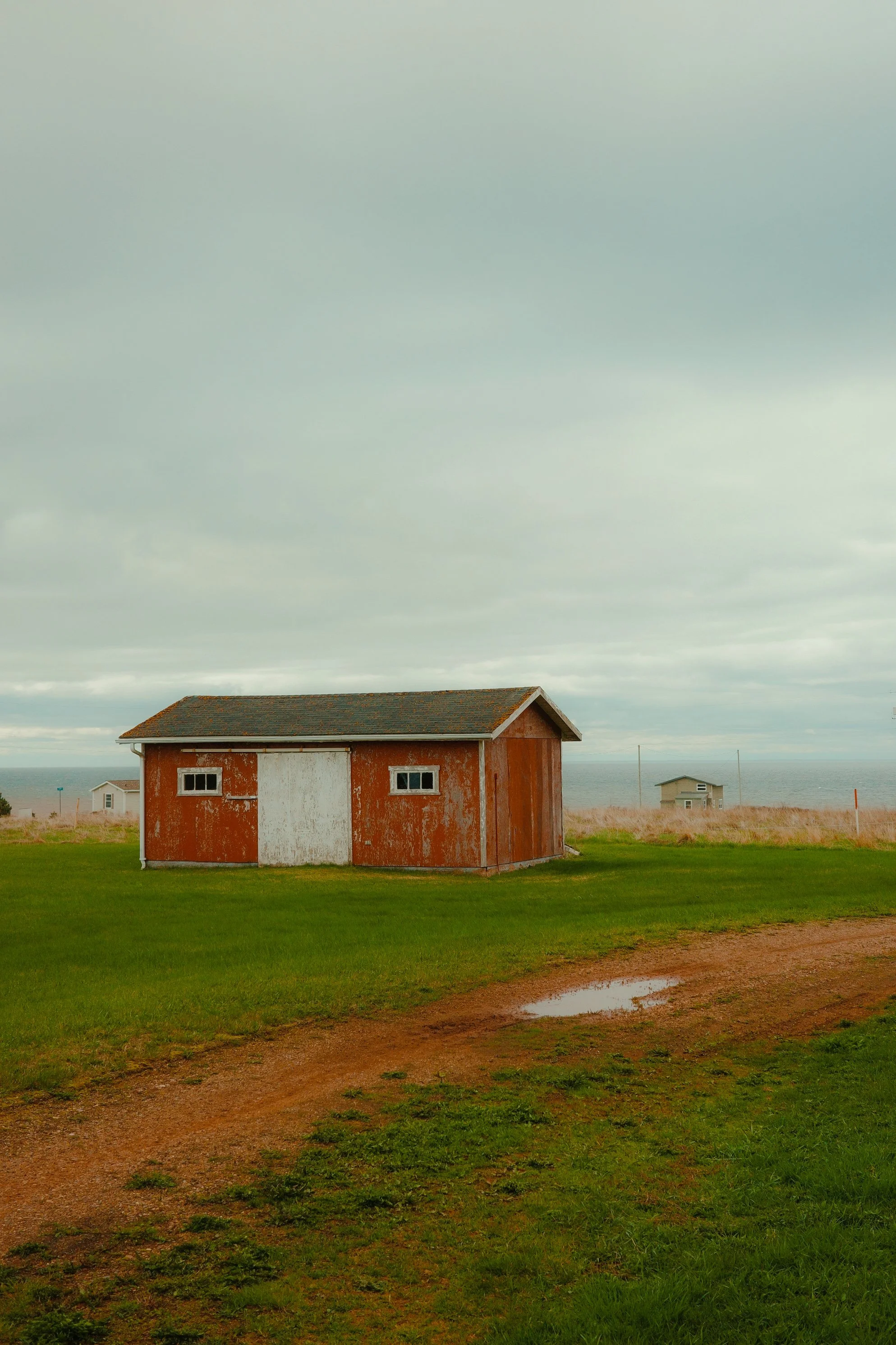 Une petite cabane en bois rouge et blanc située dans un champ vert, avec un chemin de terre boueux et une mare d'eau, et des maisons en arrière-plan près de la mer sous un ciel nuageux.