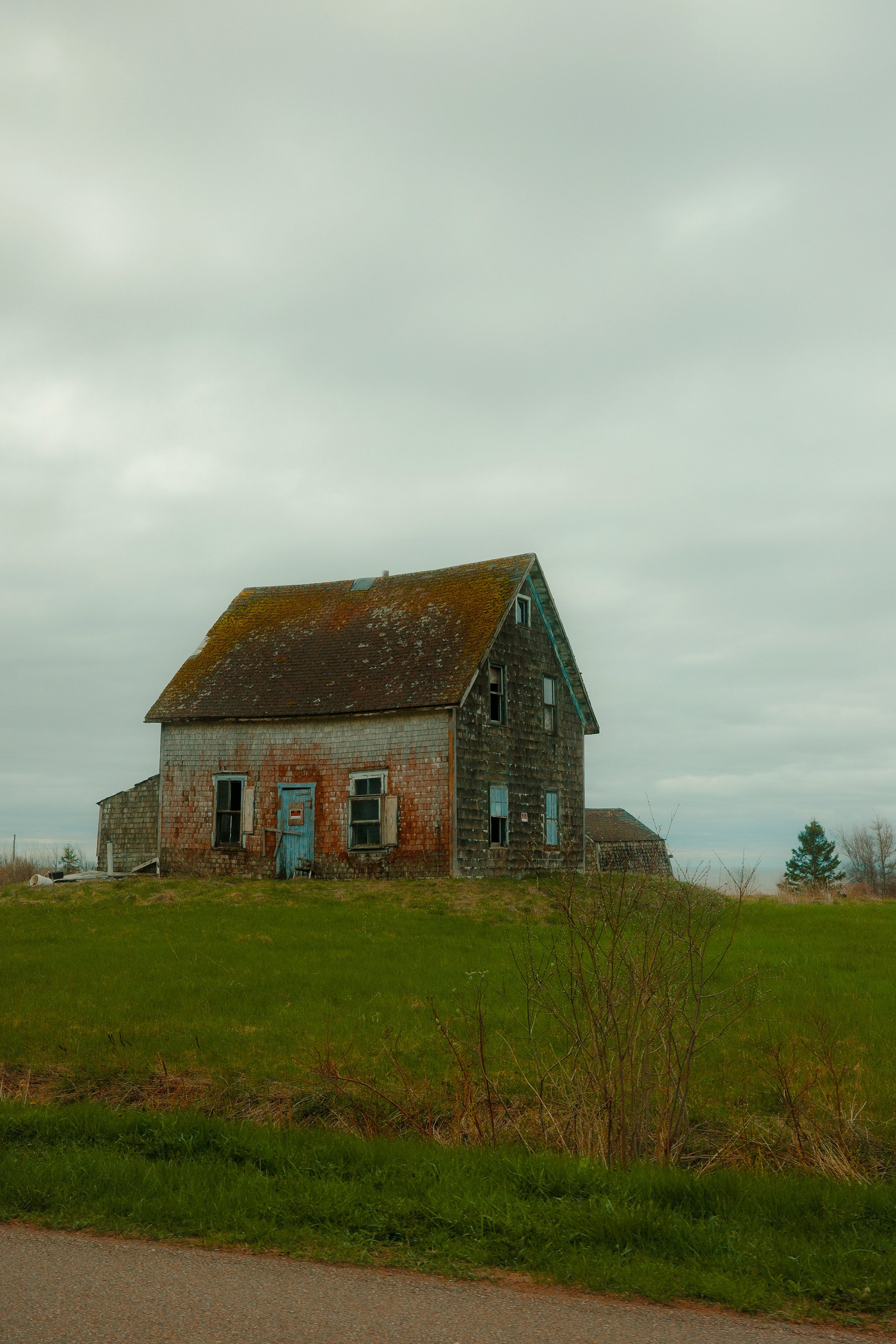 Une maison abandonnée en bois avec un toit en tuiles, entourée d'herbe verte et un ciel nuageux.
