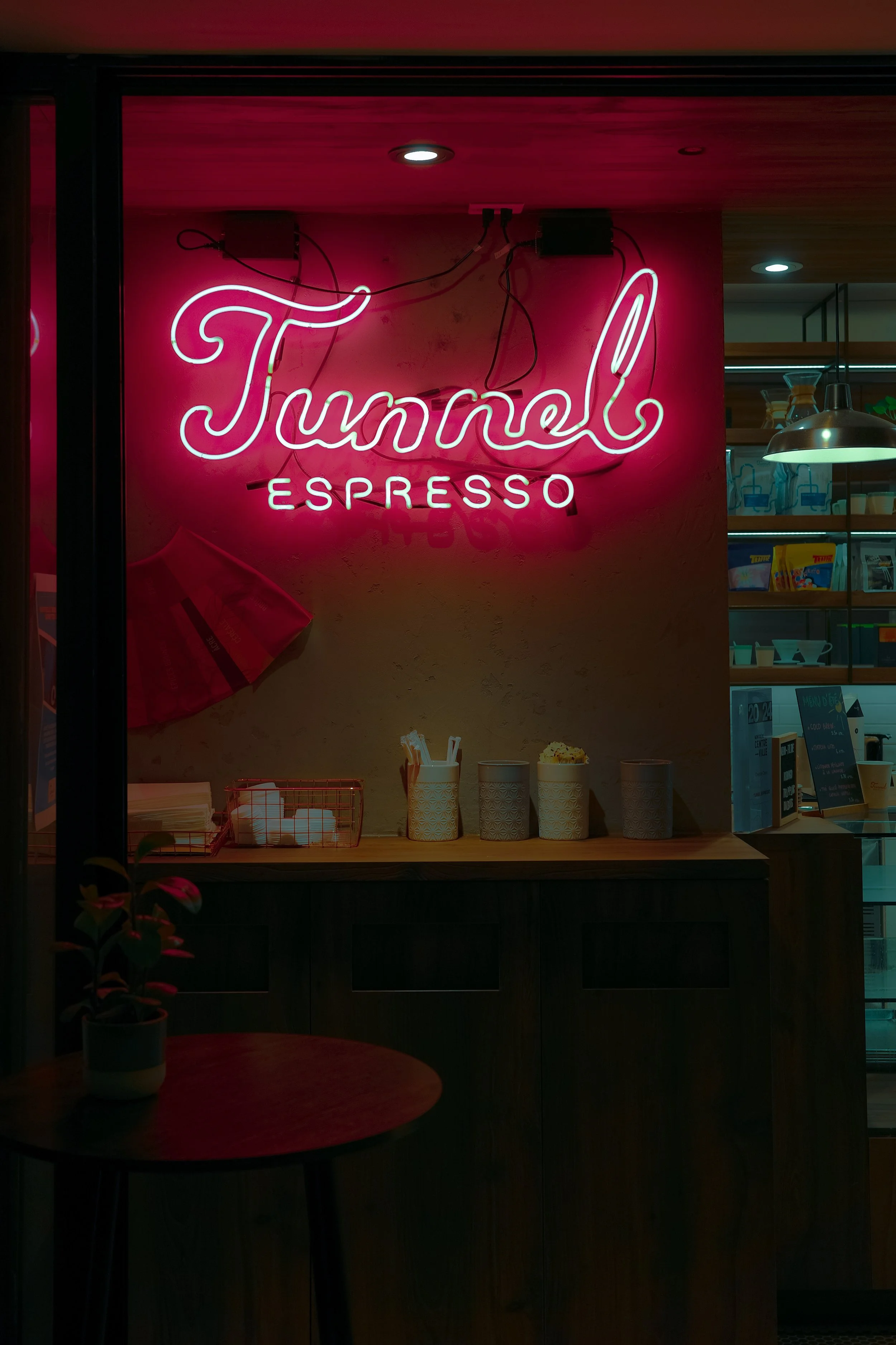 Un panneau lumineux rose avec l'inscription 'Tunnell ESPRESSO' à l'intérieur d'un café ou d'une boutique. Il y a un petit comptoir avec des gobelets et des ustensiles, et une étagère avec des produits ou des machines derrière.