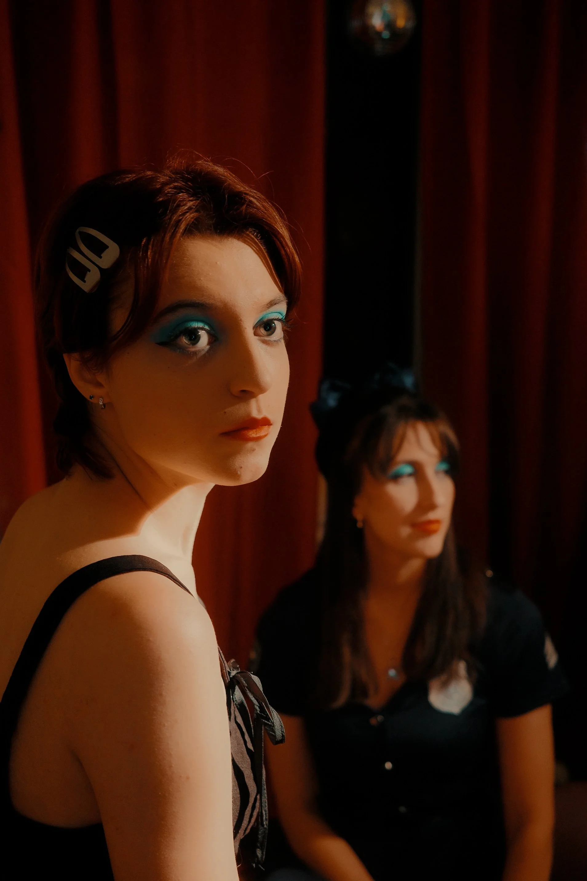 Deux jeunes femmes avec maquillage bleu, assises devant un rideau rouge, regardant loin.