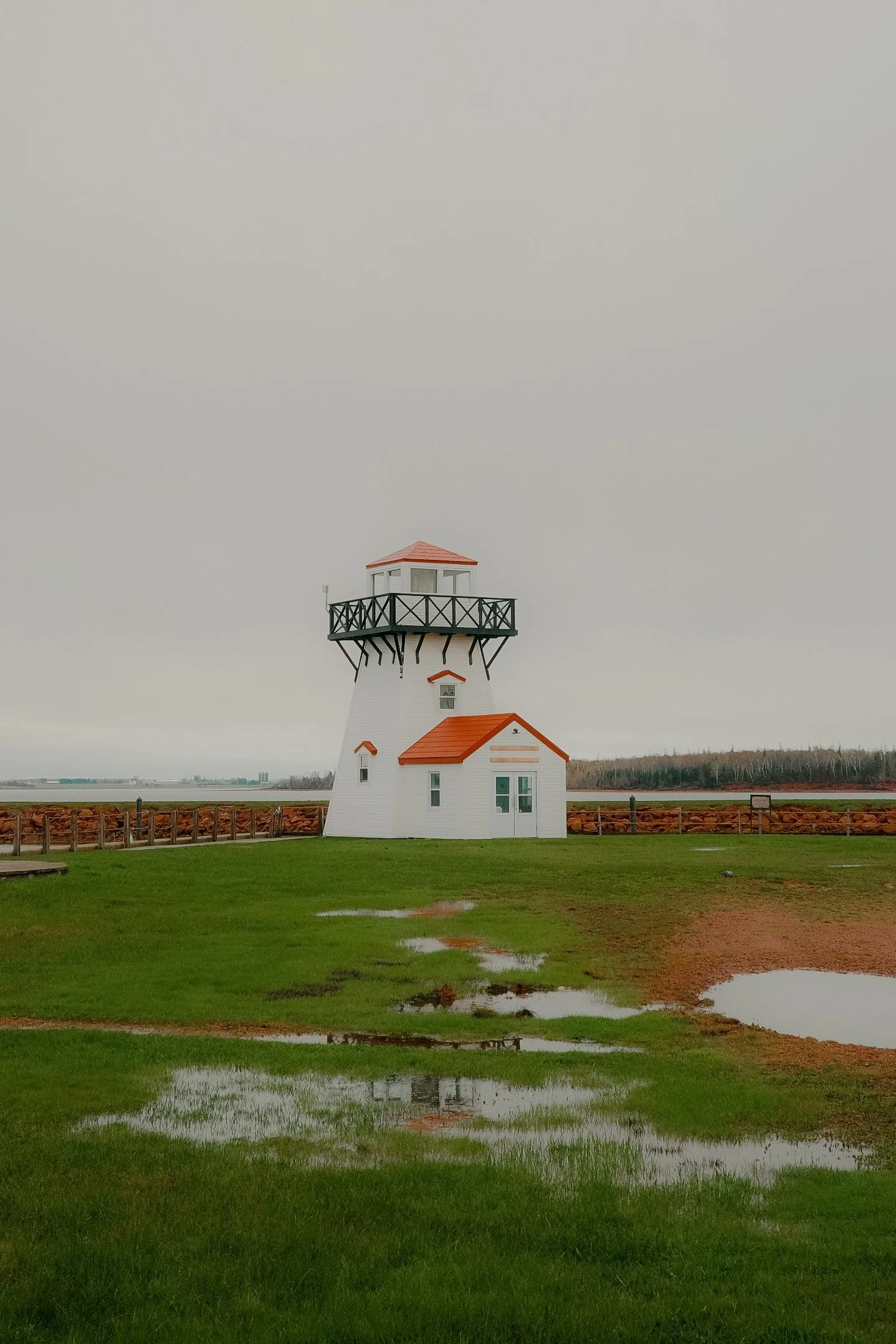 Un phare blanc avec une toiture rouge, situé sur un terrain herbeux avec des flaques d'eau et entouré d'une clôture en bois, sous un ciel gris et nuageux.