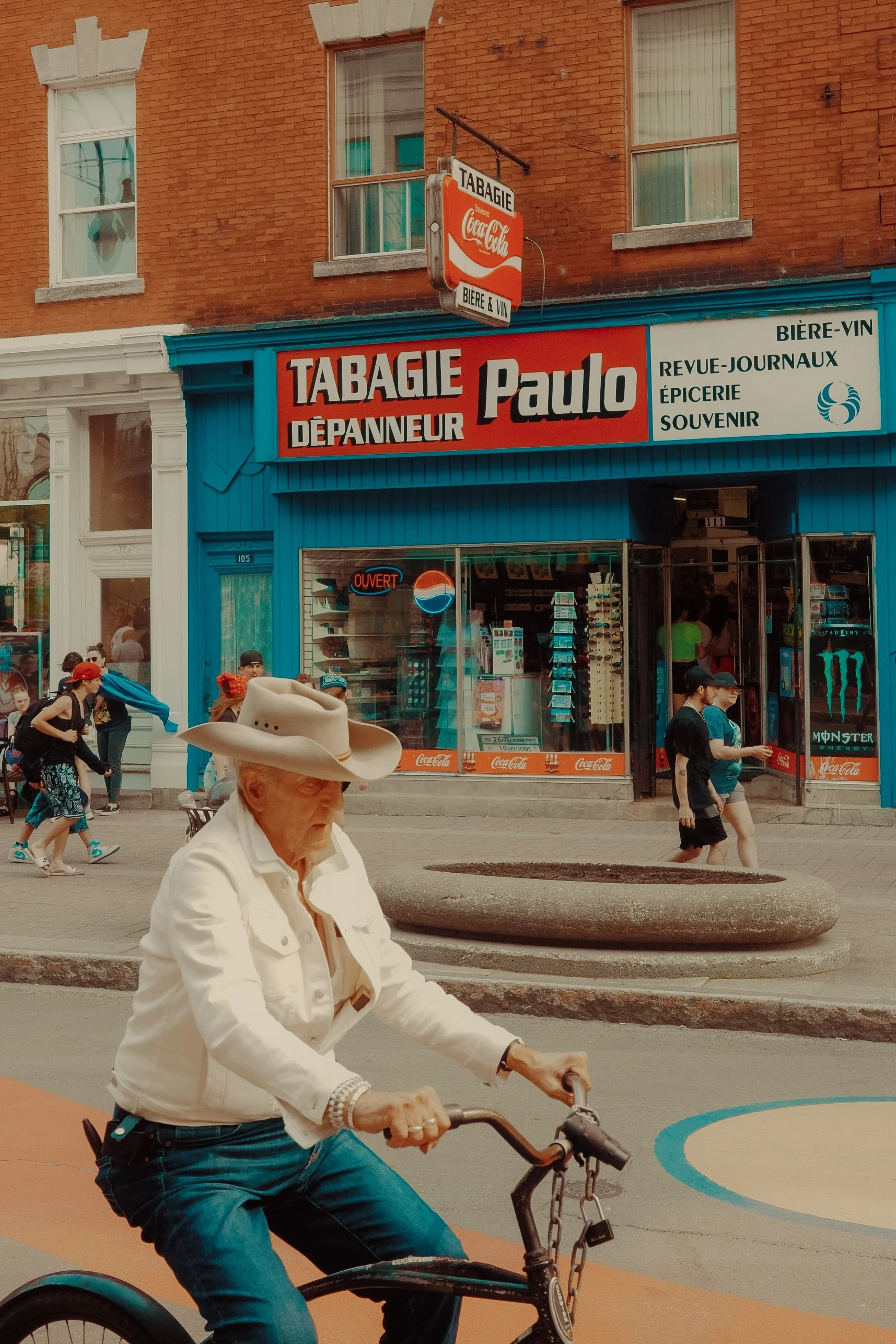 Un homme âgé portant un chapeau cowboy blanc et une veste blanche, qui fait du vélo dans une rue animée, avec un magasin en arrière-plan portant une enseigne rouge et blanche 'Tabagie Paulo'.