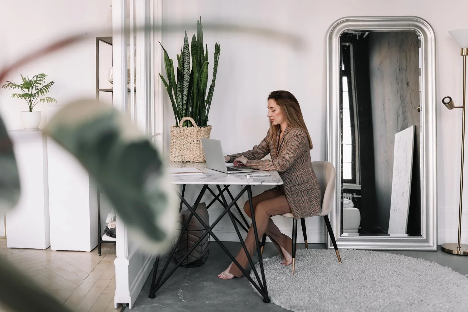 Woman working on on the SEO of her website at laptop at a desk in a modern, well-lit room with plants and a large mirror.