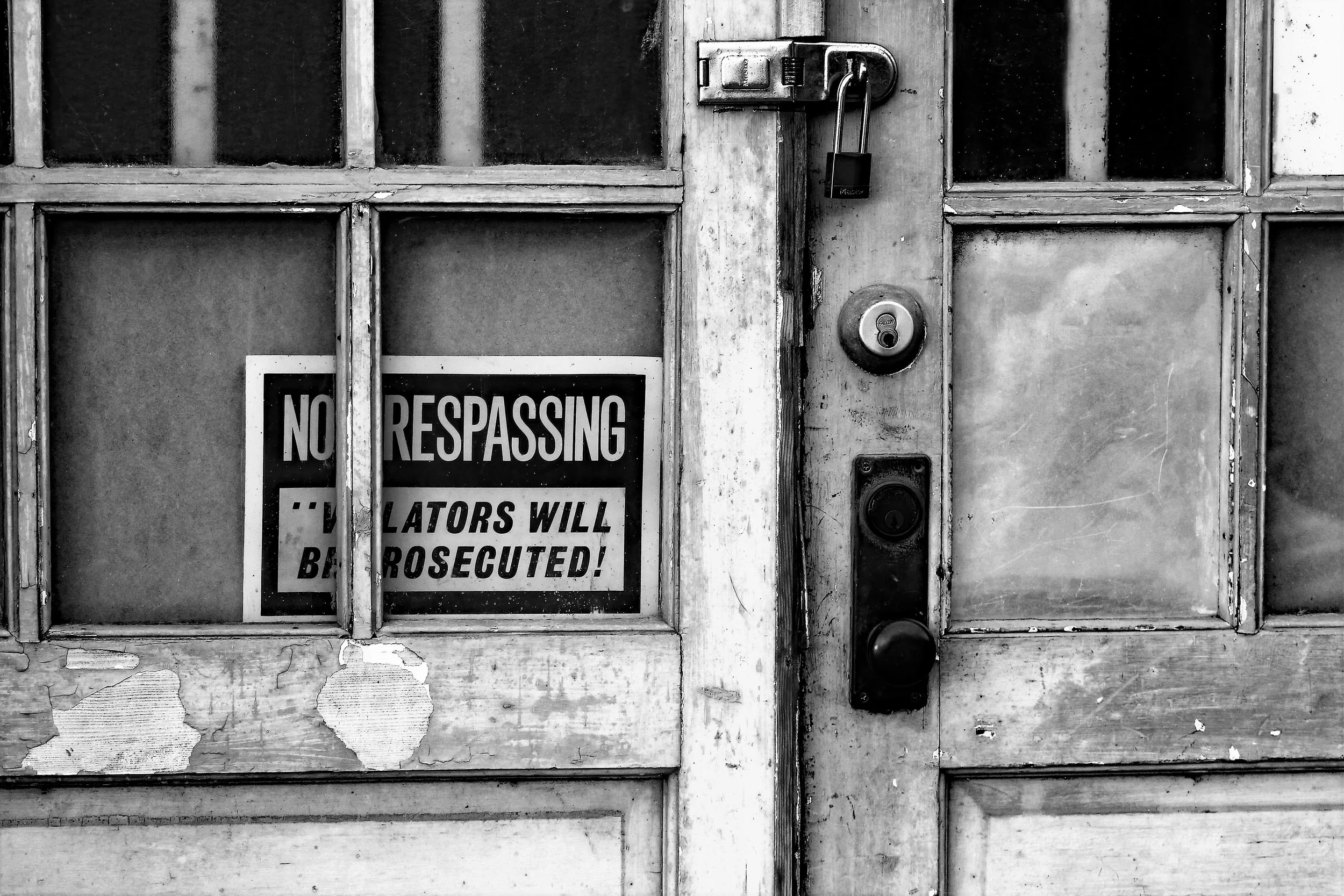 Black and white photo of an old, worn wooden door with multiple glass panes and a lock. A sign on the lower left reads 'No Trespassing. Violators will be prosecuted.' The door has peeling paint and visible weathering.