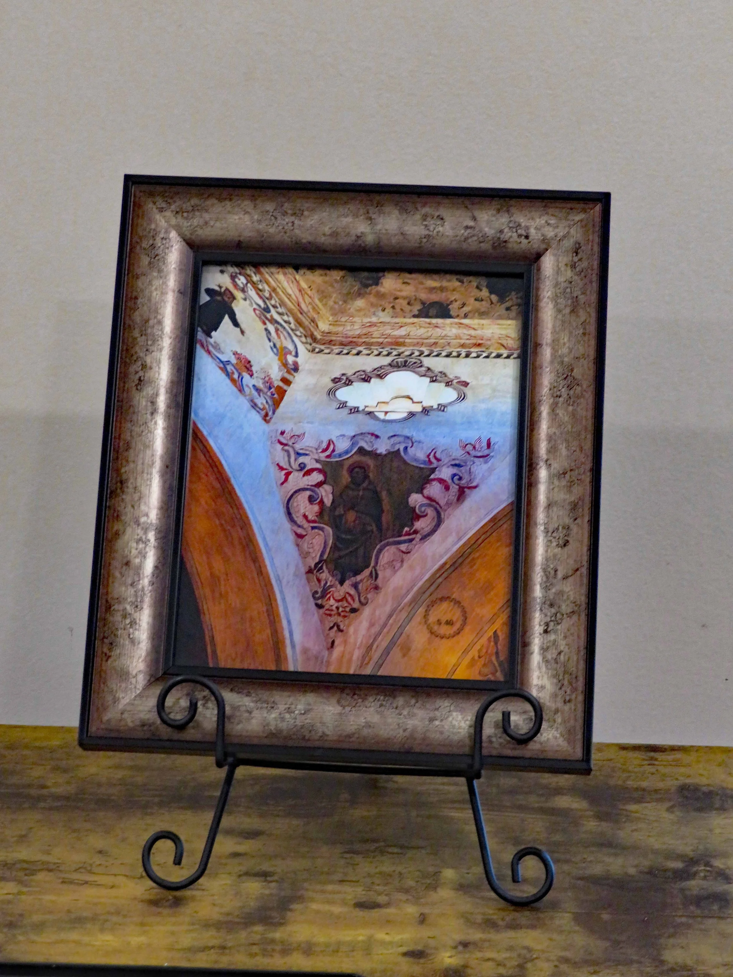 Framed mirror with ornate painted ceiling reflection and wall, placed on a wooden table with metal scroll legs.