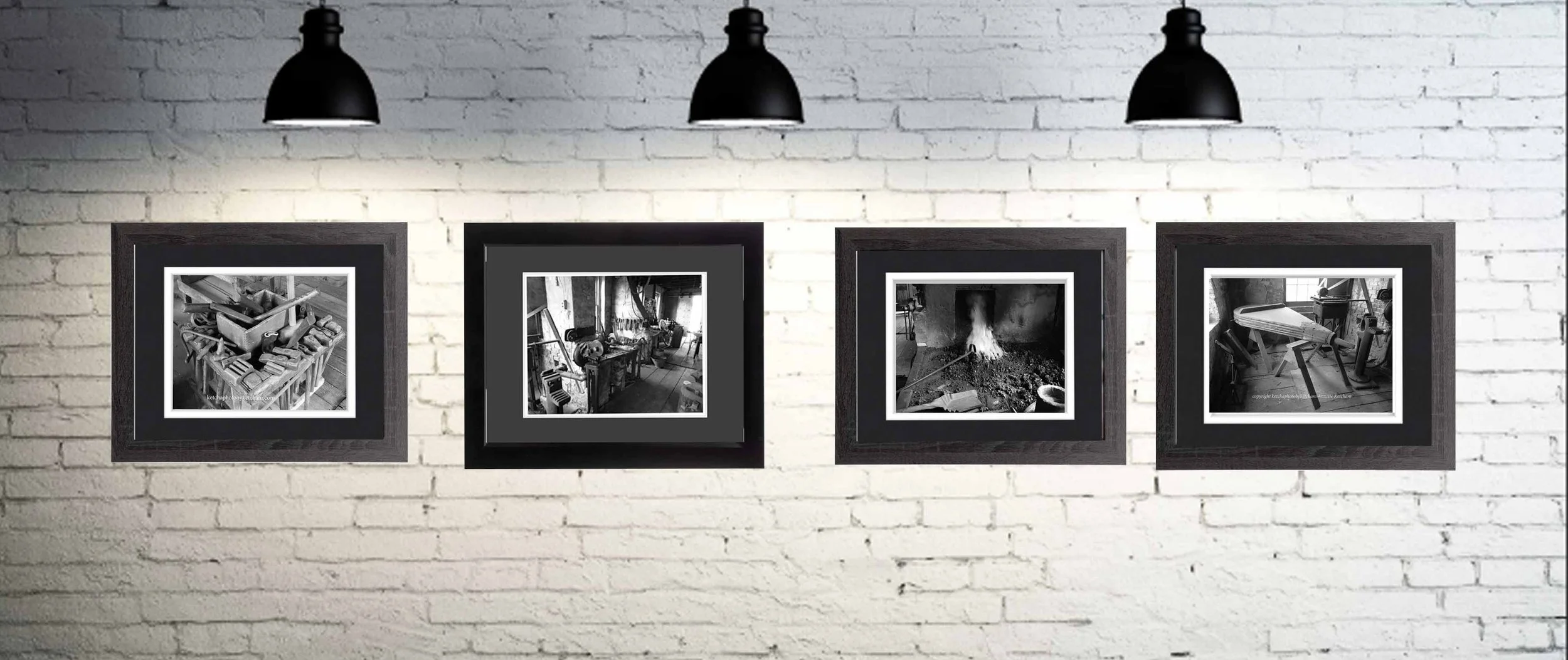 Four black-and-white framed photographs hung on a white brick wall, illuminated by black ceiling lamps above.