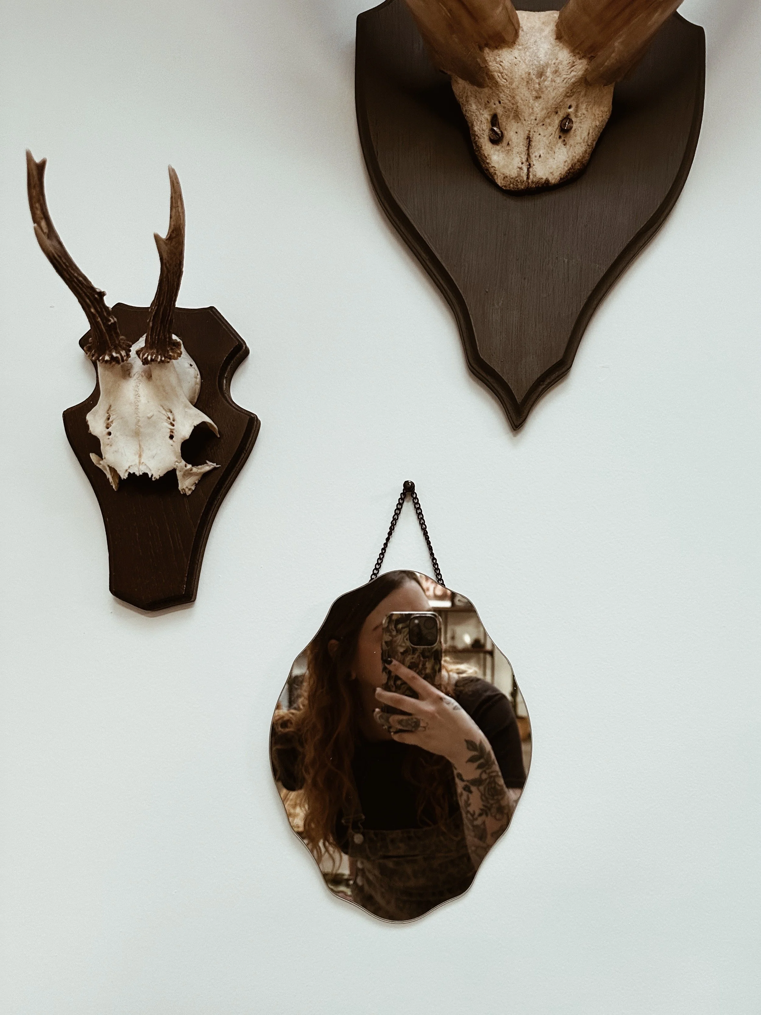 Een muur met twee houten dierenhoofden en een spiegel waarvan de rand de vorm heeft van een vogel. In de spiegel is een vrouw met rood haar zichtbaar die een telefoon vasthoudt.