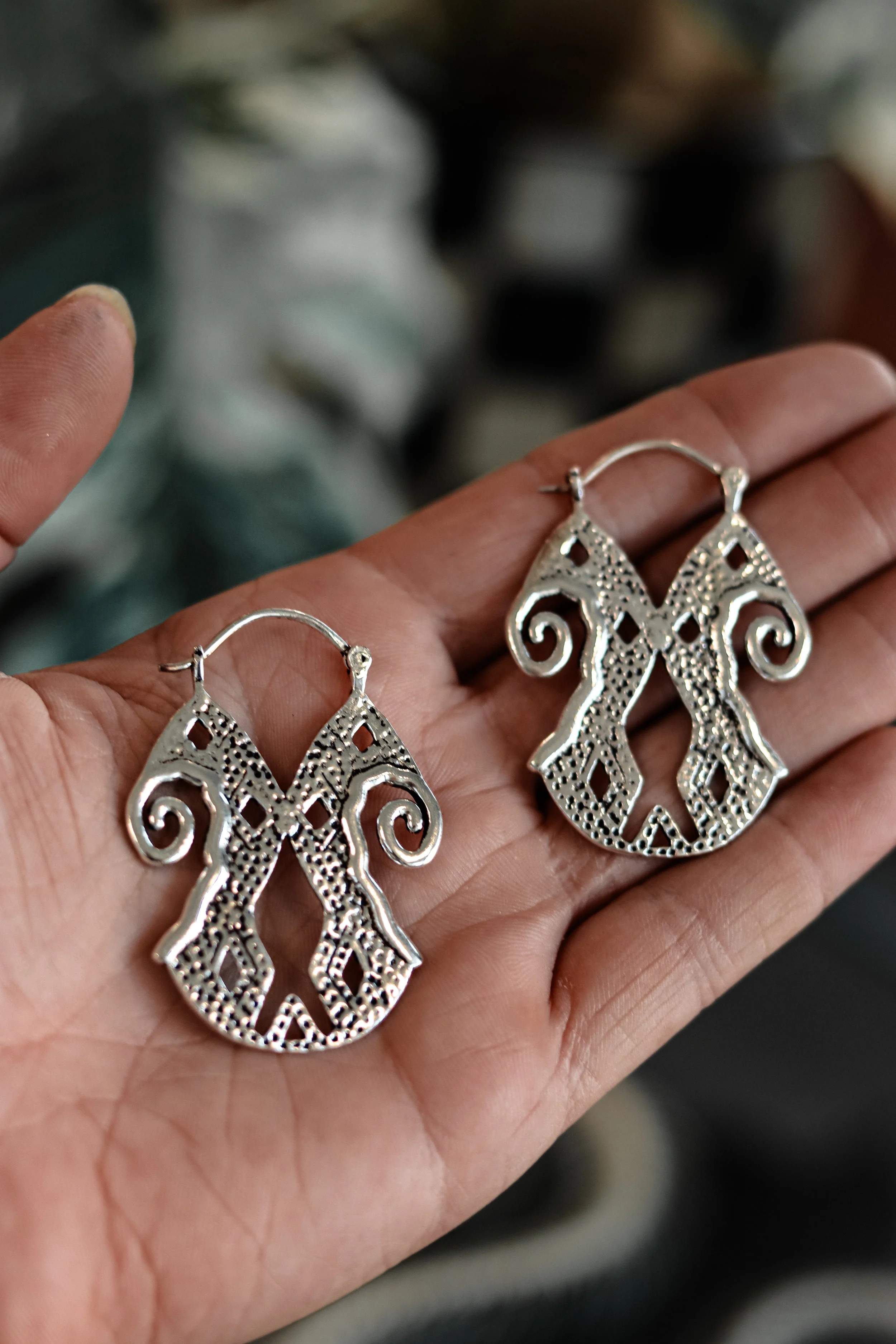 Een hand die een paar zilveren oorbellen vasthoudt met een vakmanschap ontwerp, met een achtergrond van een ietwat onscherpe omgeving.