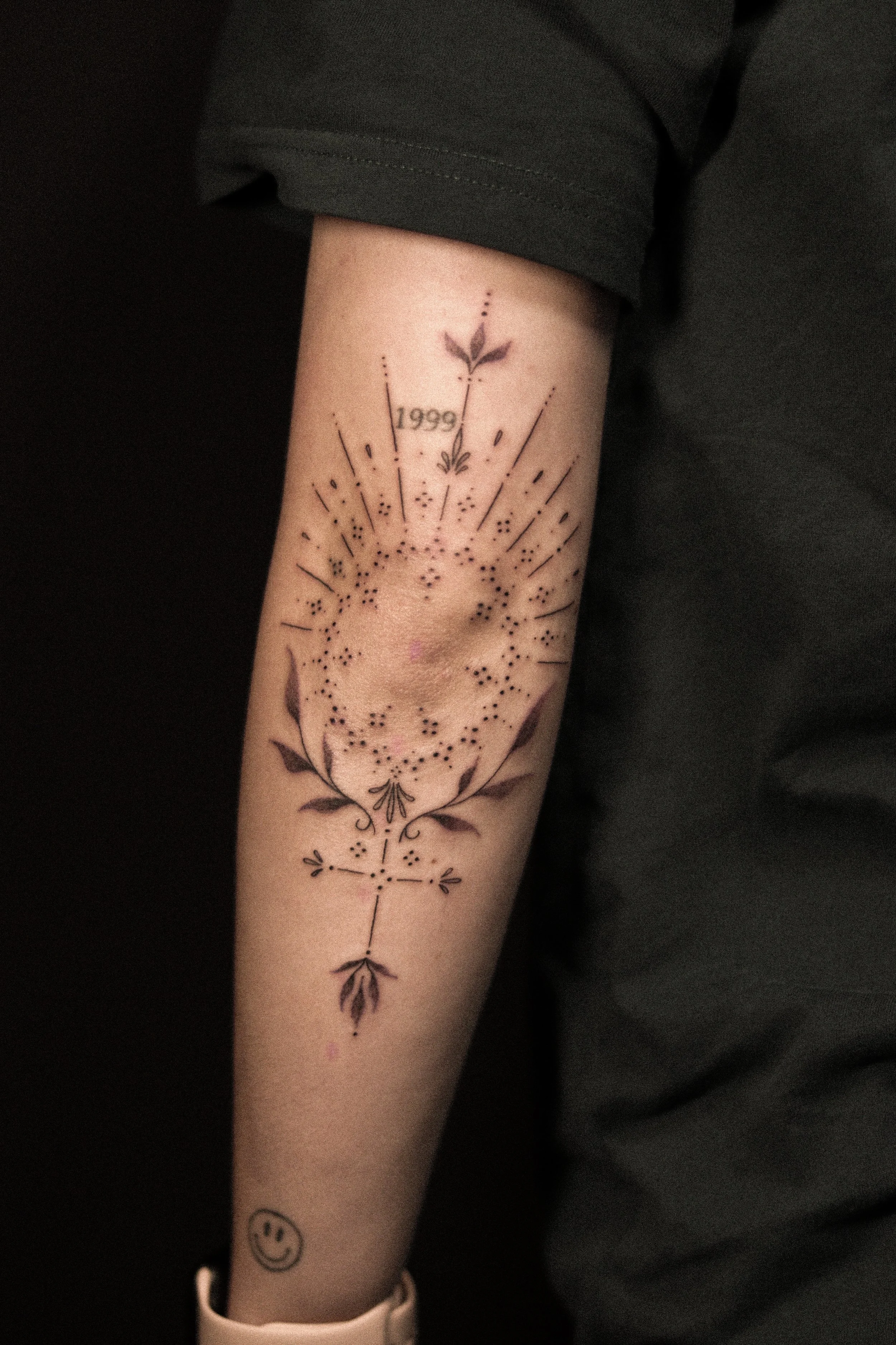 Tatoeage op onderarm met datum 1999, bladeren, kleine bloemen en een lachend gezicht.
