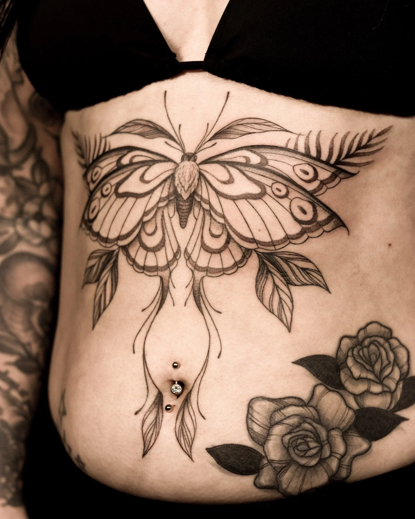 Tattoo van een grote vlinder met bladeren en bloem op de buik, plus piercings in de navel.