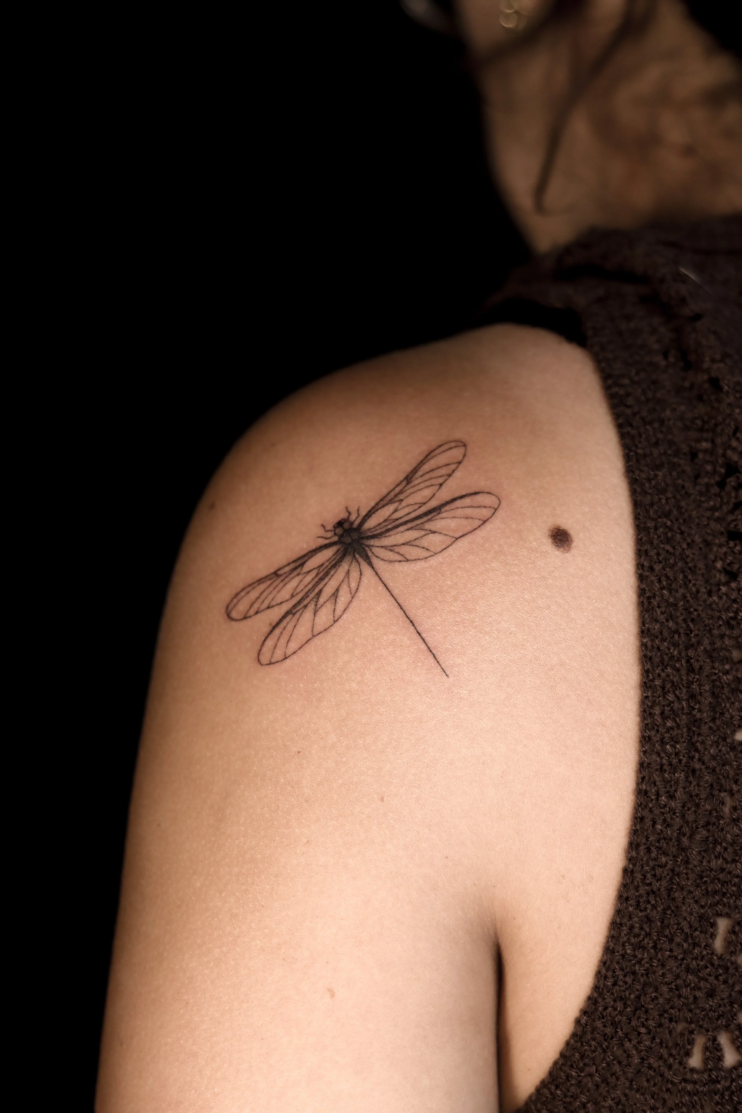 Tattoo van een libelle op de schouder met een kleine moedervlek ernaast.