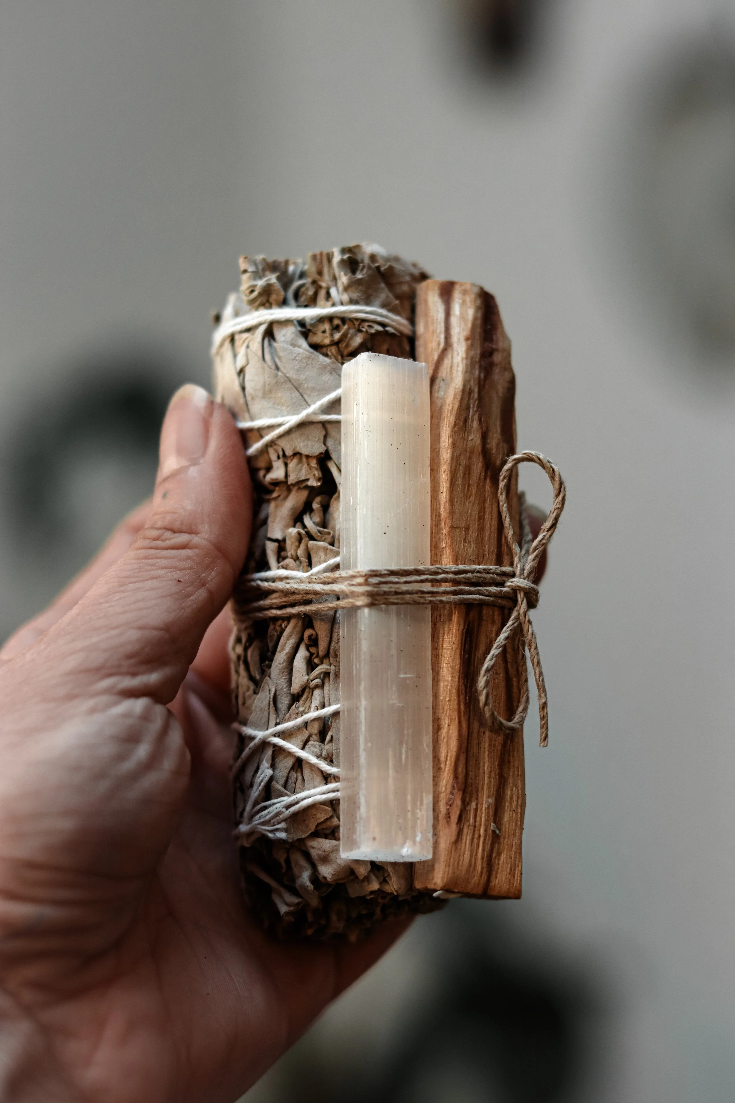 Hand die een bundel gedroogde kruiden vasthoudt, met een stukje hout en een kristal, allemaal stevig samengebonden met touw.