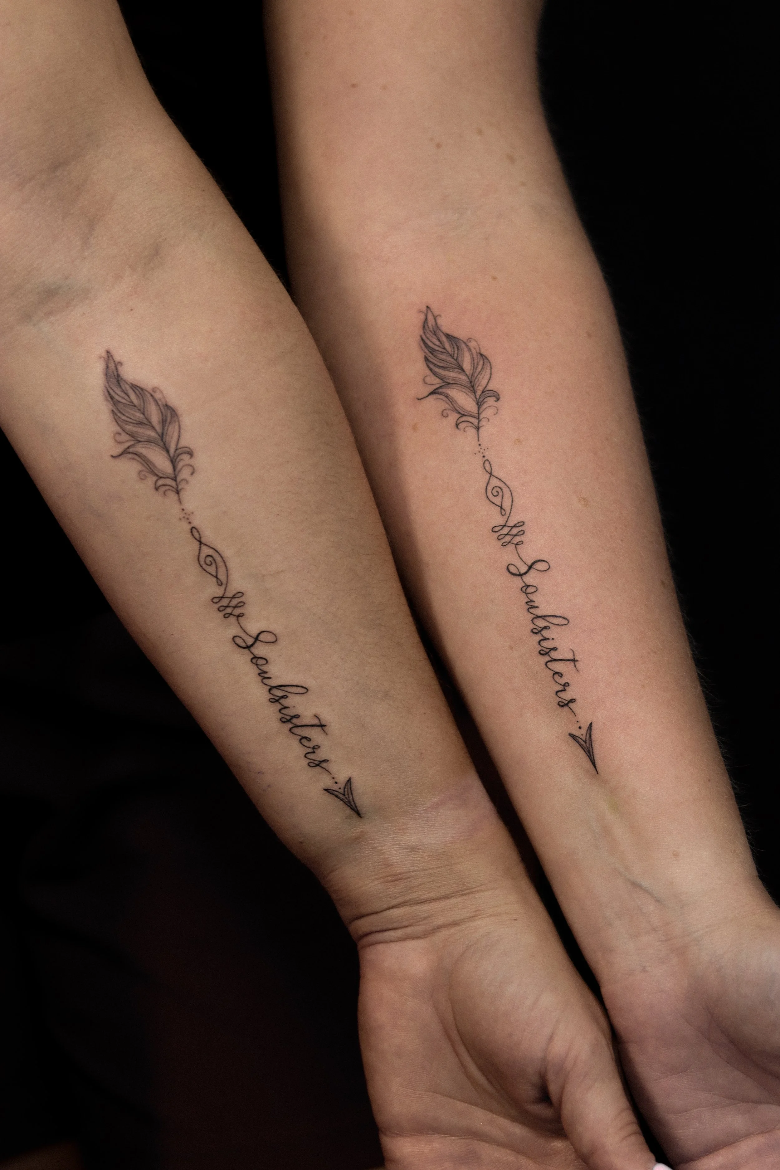 Twee onderarmen met identieke tatoeages van een veer en cursief schrift dat zegt 'The Sandflutterers'.