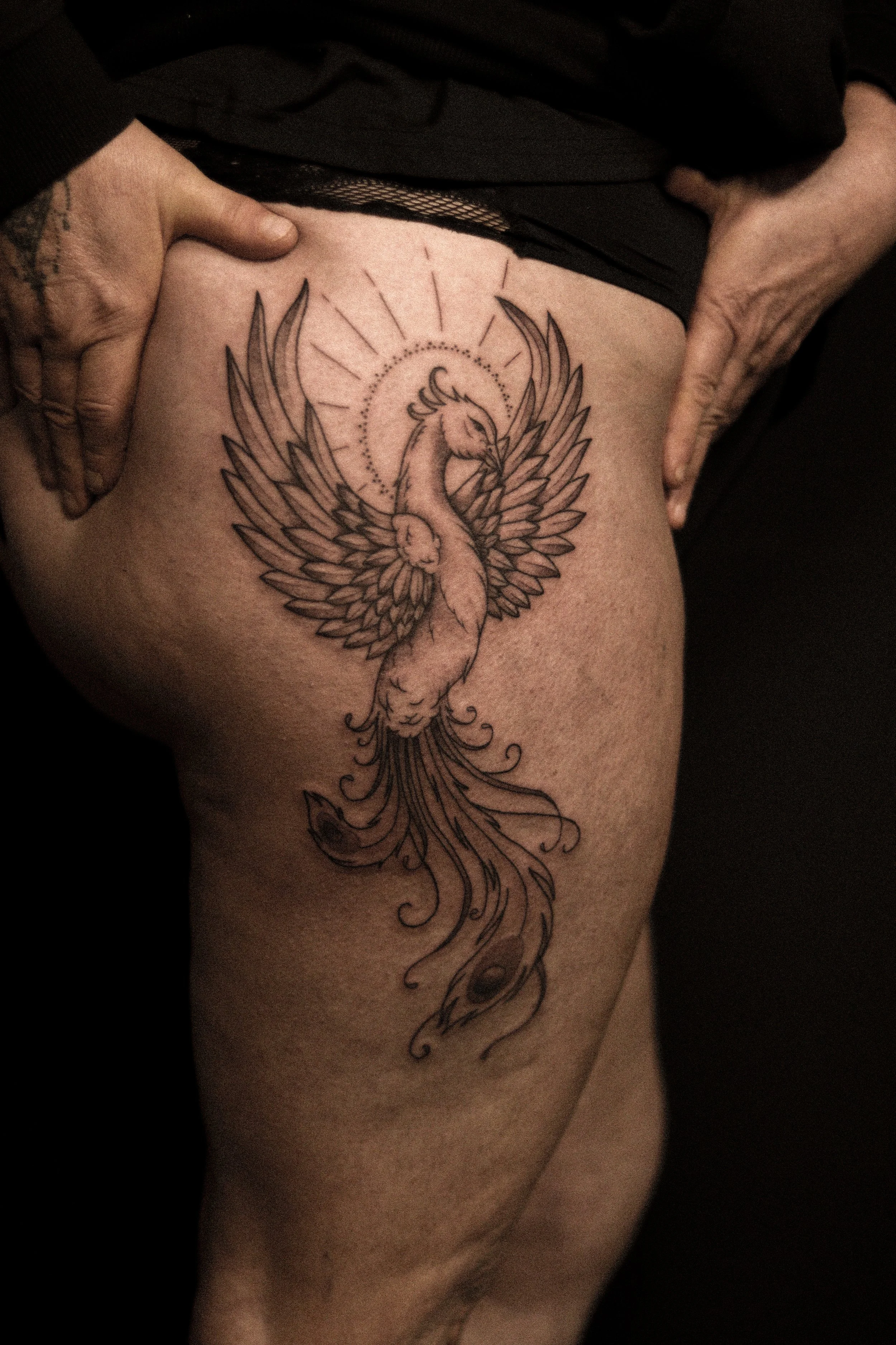 Tatoeage van een feniks met uitgebreide vleugels en een lange, sierlijke staart, geplaatst op het dijbeen.