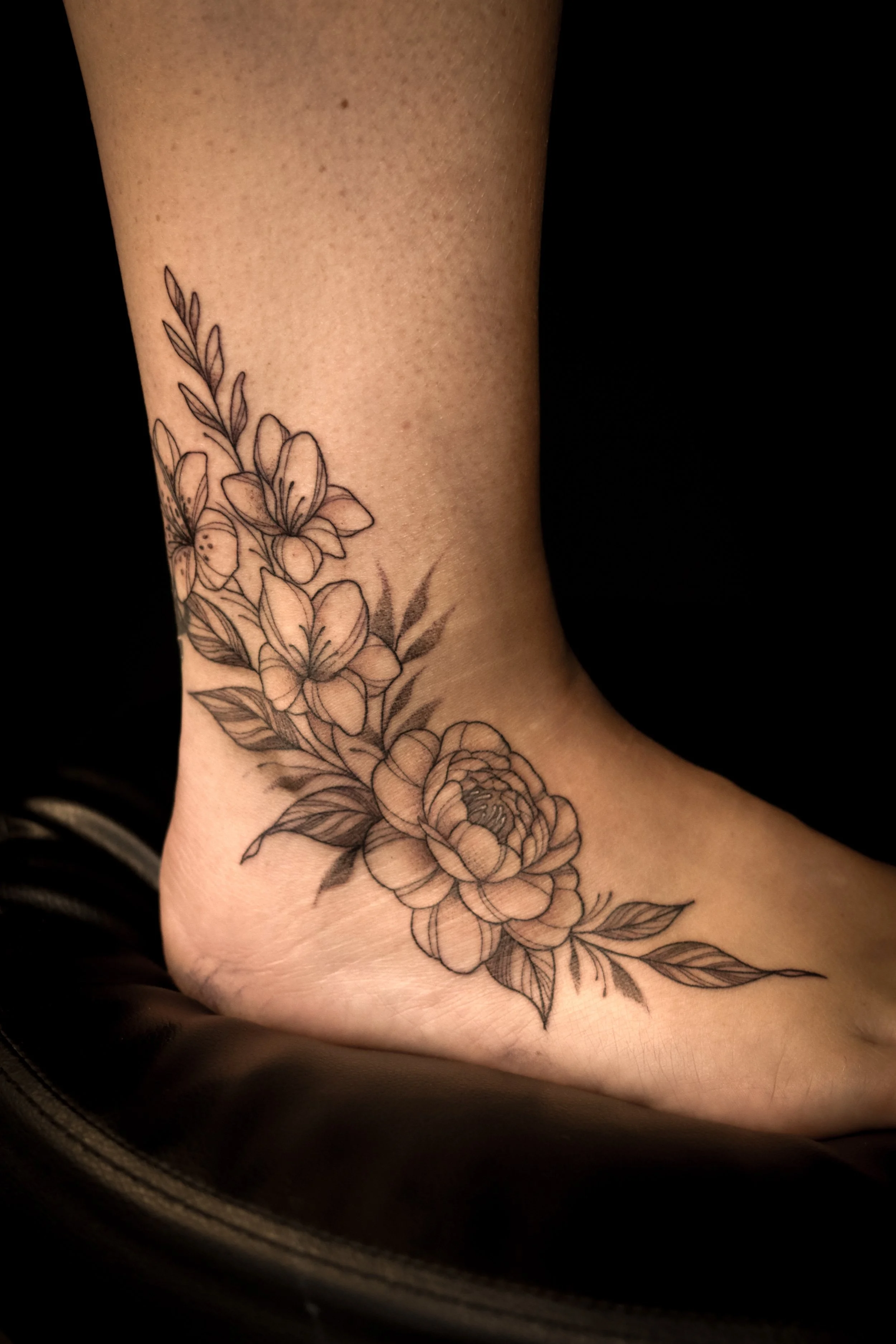 Een enkelvoudige zwarte lijn-tatoeage van bloemen en bladeren op de enkel en voet.