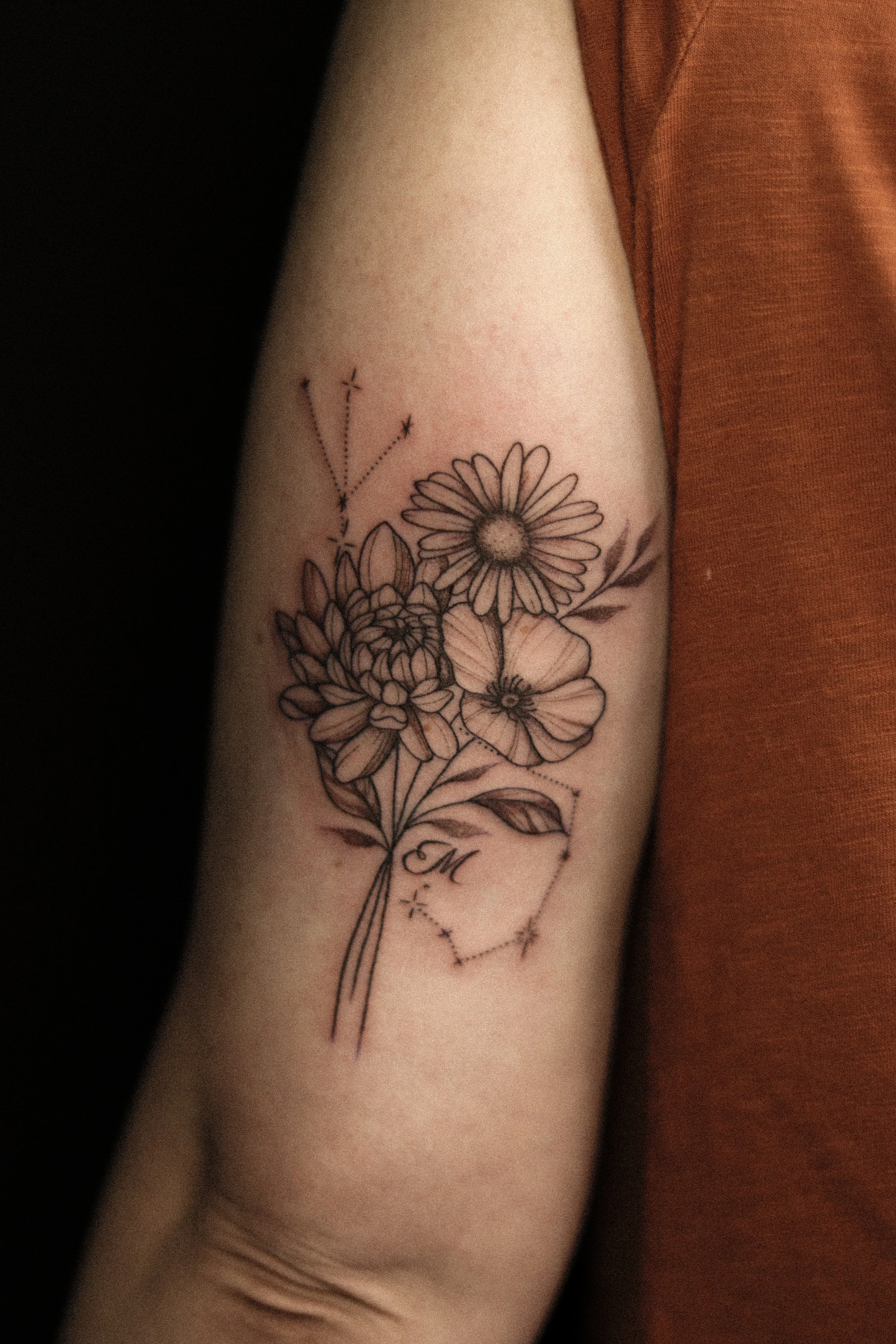 Tatoeage van bloemen met een letter 'M' en een sterrenbeeld op de bovenarm.