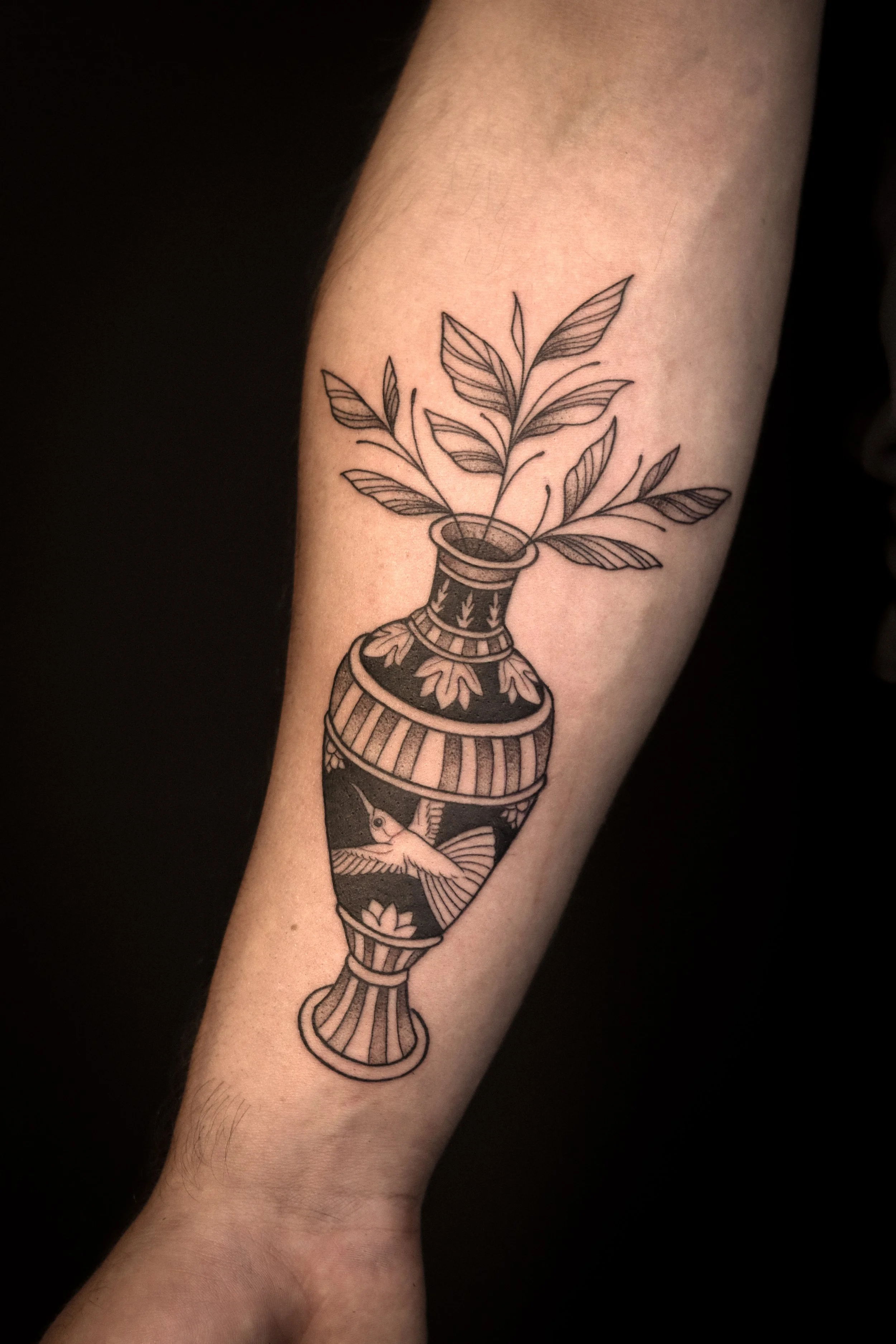 Tatoeage van een sierpot met bloemen en een kolibri op de onderarm.