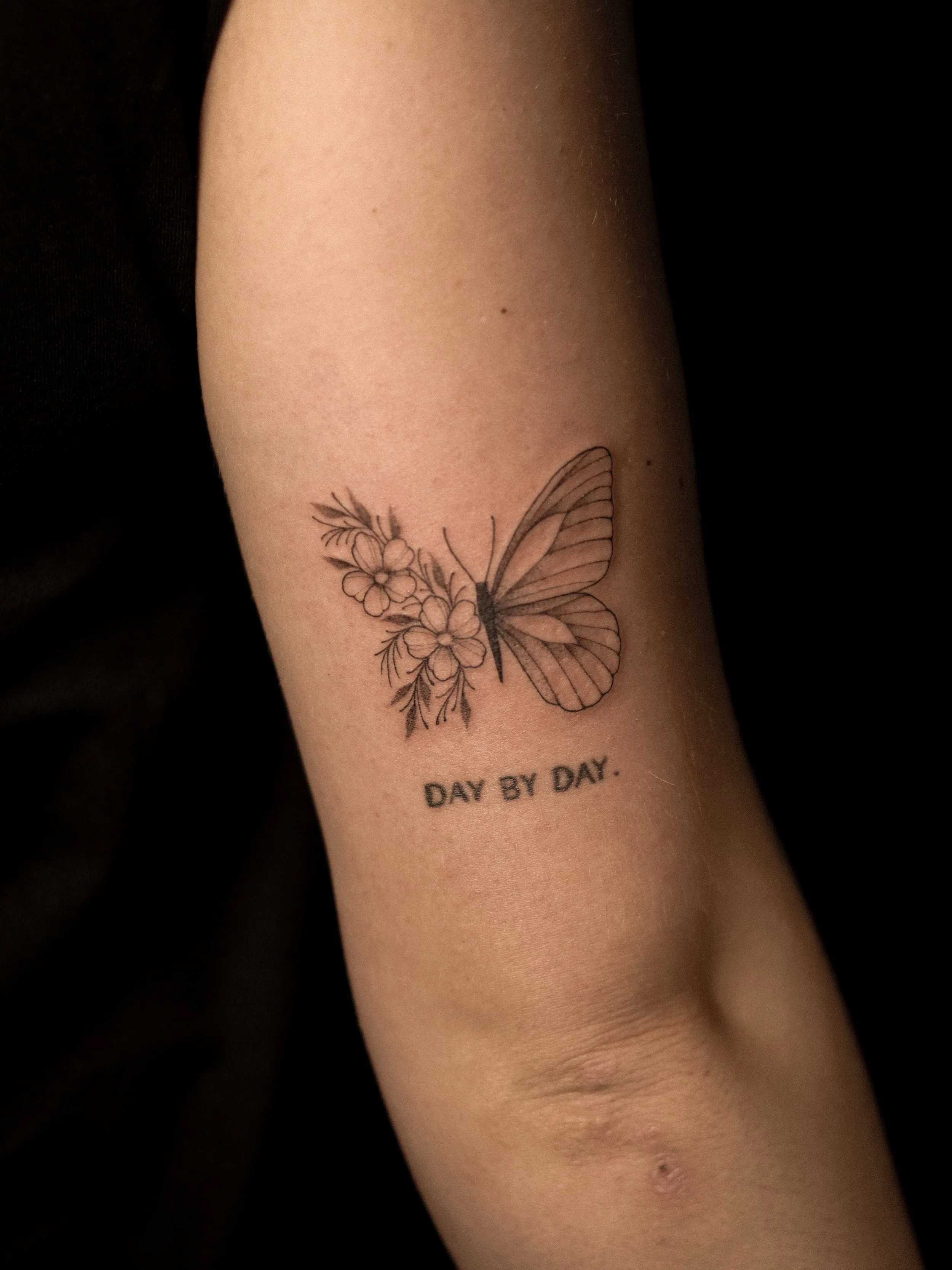 Tatoeage van een vlinder en bloemen op de onderarm met de tekst 'DAY BY DAY' eronder.
