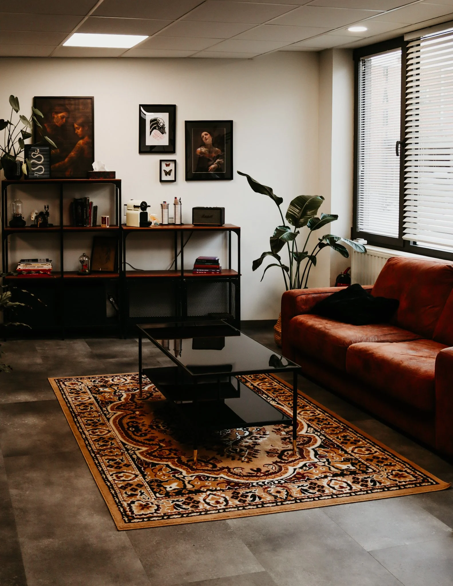 Een woonkamer met een rode bank, een zwarte salontafel, een boekenkast, wanddecoraties en planten, met een raam met luiken.